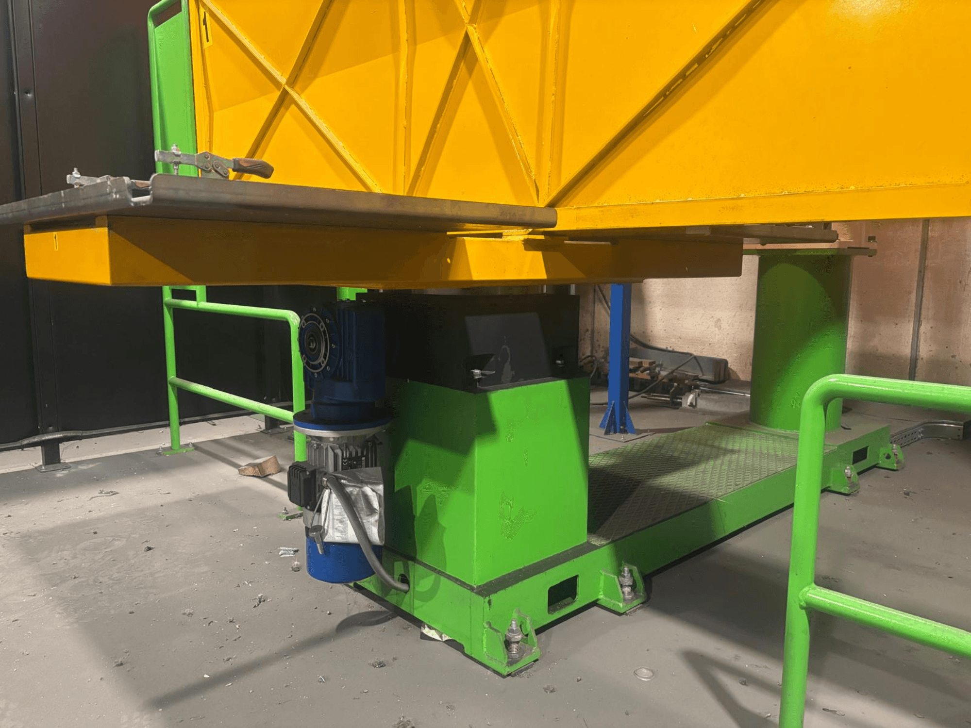 Maquinaria industrial verde e amarela numa vista frontal baixa, com um motor e uma plataforma com uma superfície texturada.