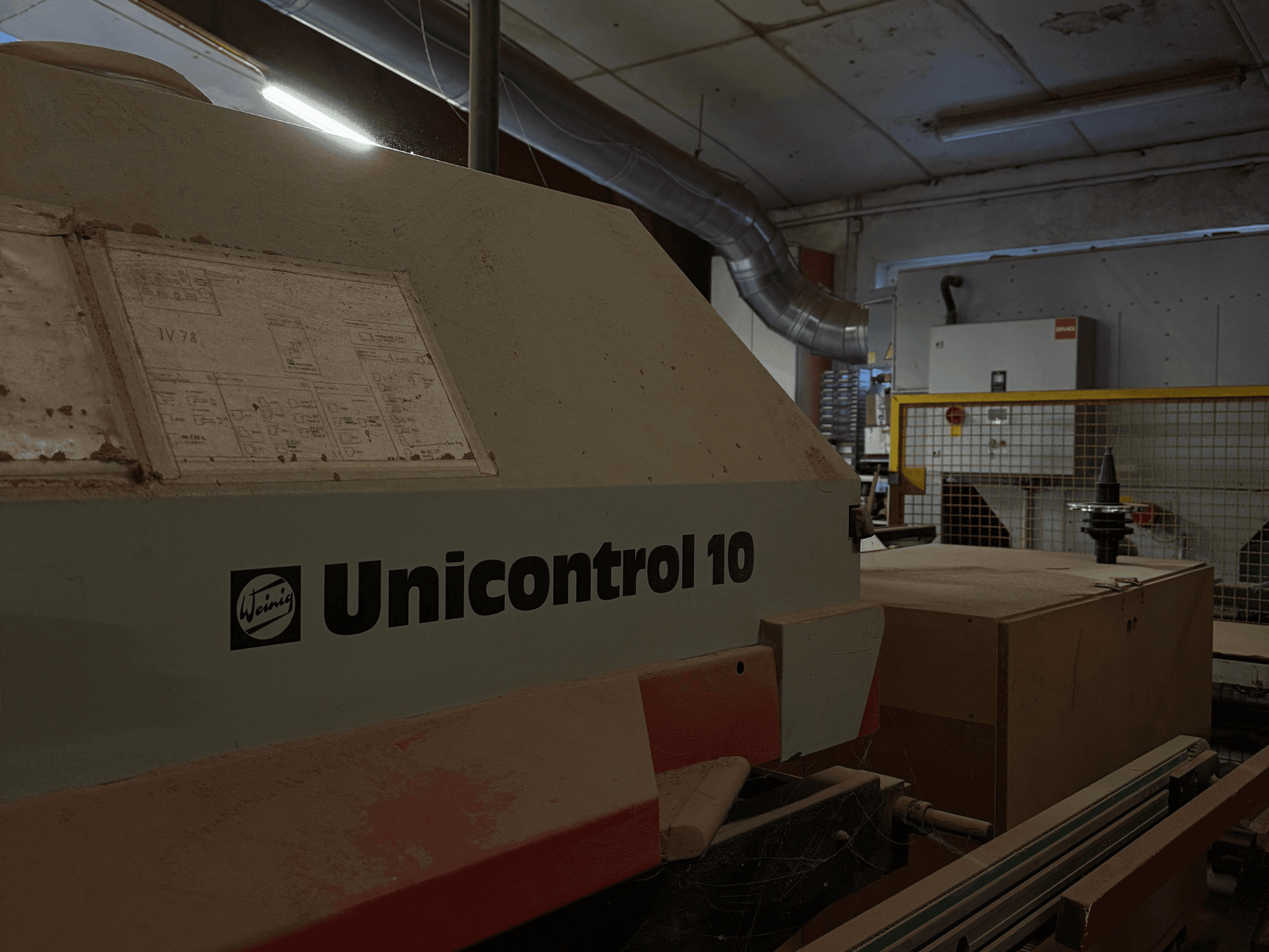 Máquina Weinig Unicontrol 10 vista de um ângulo lateral, superfície poeirenta, esquemas técnicos visíveis no painel frontal.