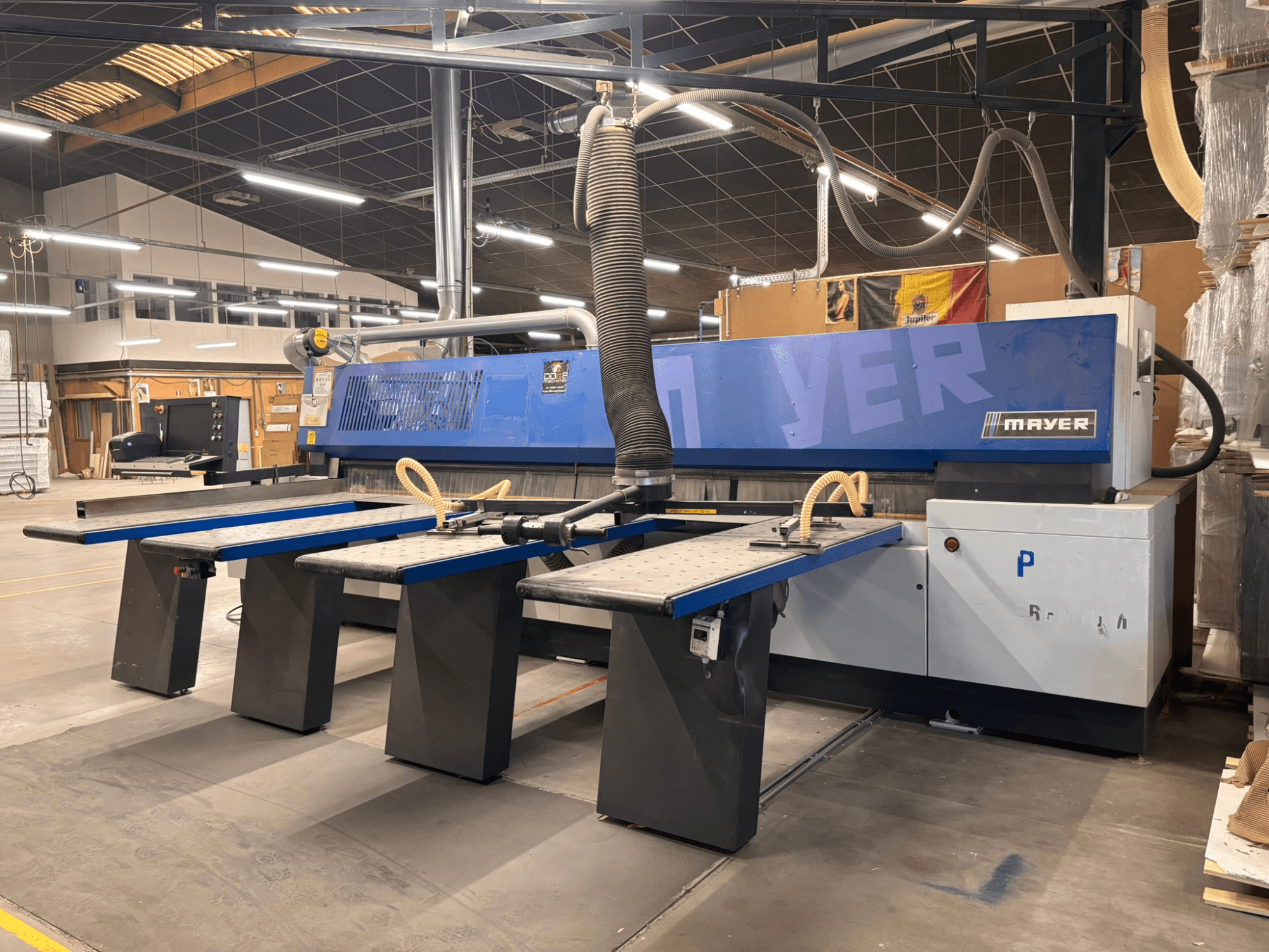 Máquina CNC Mayer com painéis azuis, vista frontal; possui várias mesas de trabalho e um sistema de extração de poeiras para trabalhos em madeira.