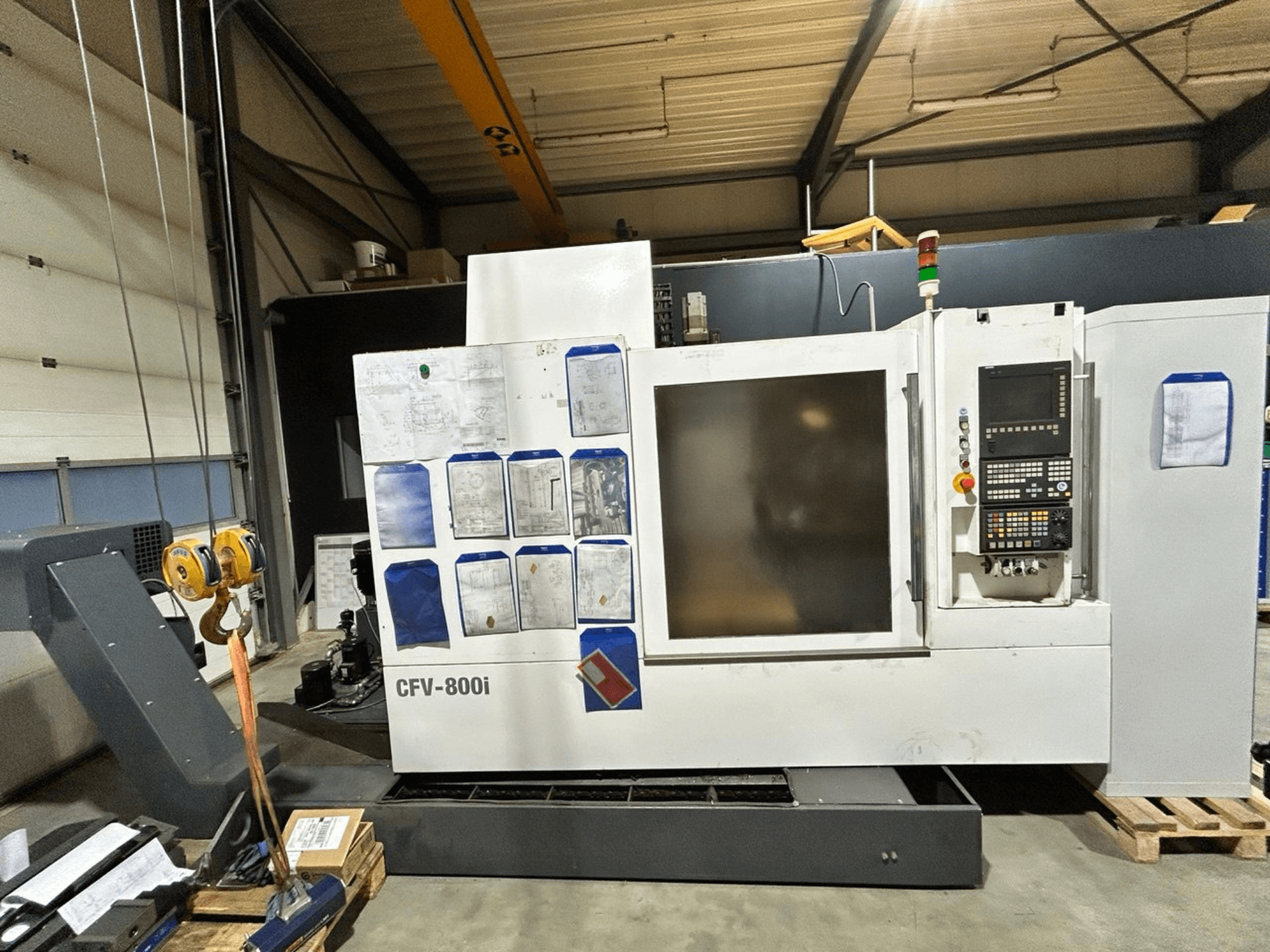 Máquina CNC CFV-800i mostrada em vista frontal, com painel de controlo, documentos ao lado e ponte rolante em configuração de fábrica.