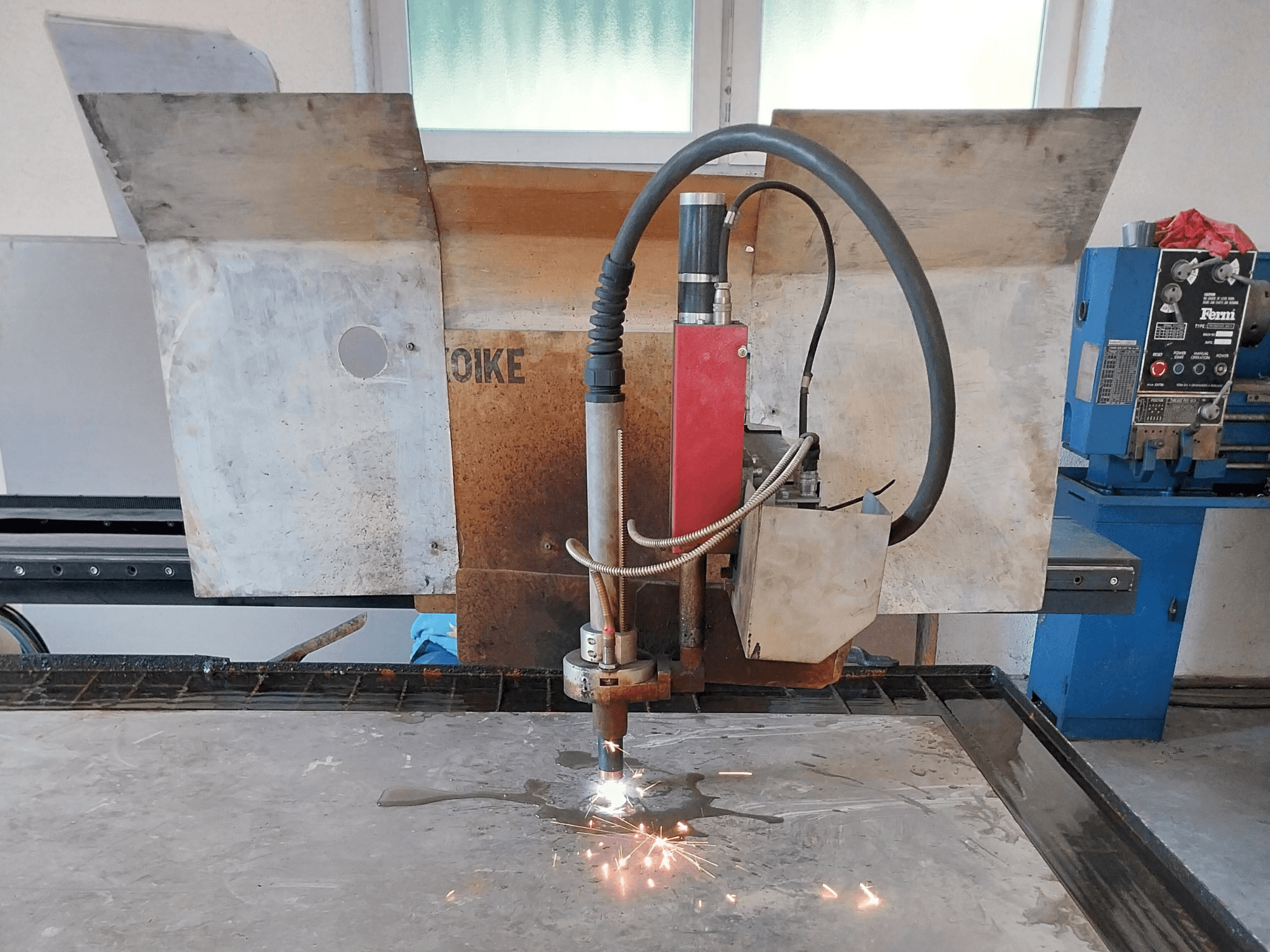 Vista frontal de um cortador de plasma Oike com faíscas a voar, posicionado sobre uma superfície metálica numa mesa de trabalho.
