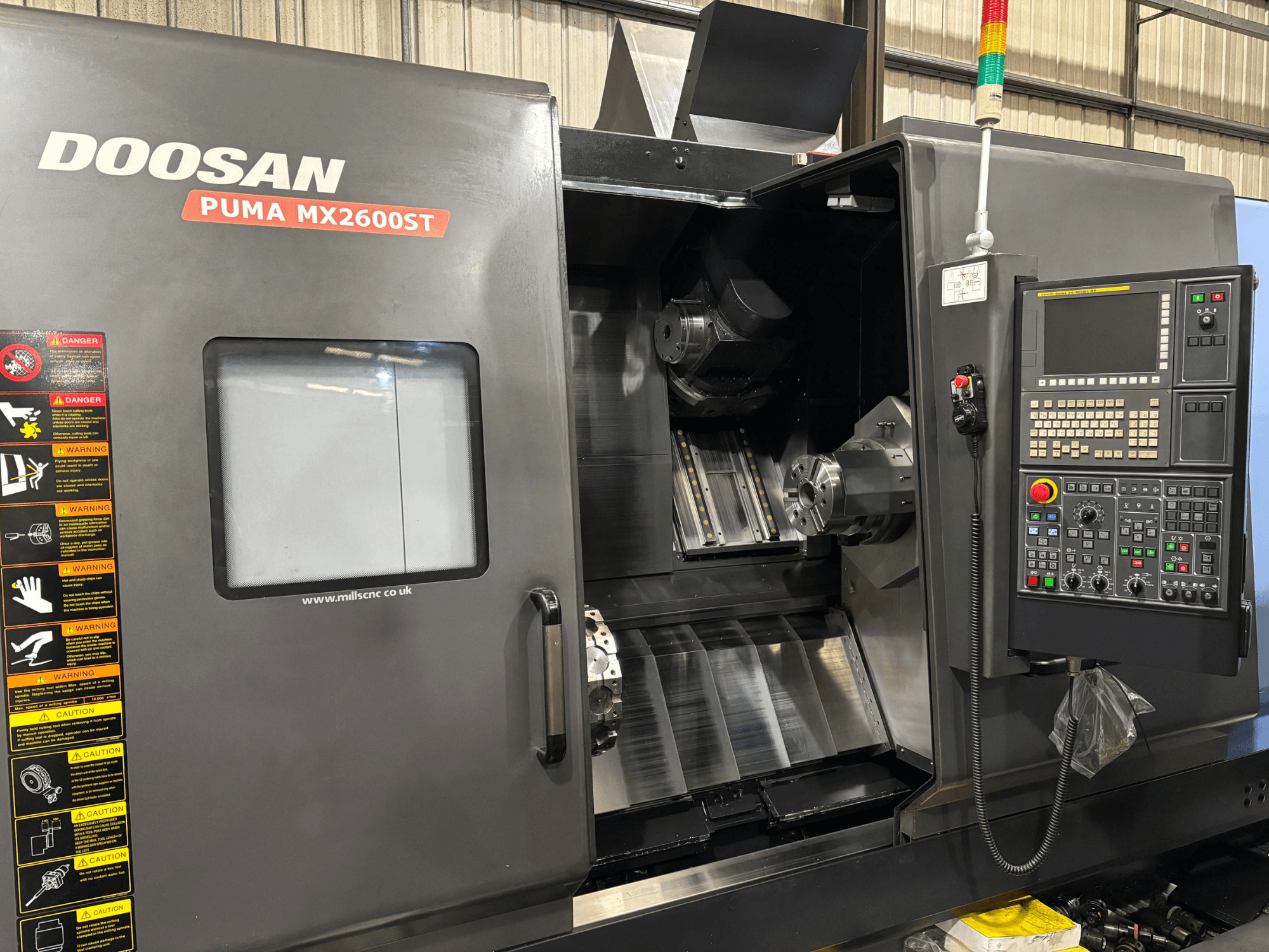 Torno CNC Doosan PUMA MX2600ST, vista frontal, mostrando o painel de controlo, o fuso e a área de maquinagem com avisos de segurança visíveis.
