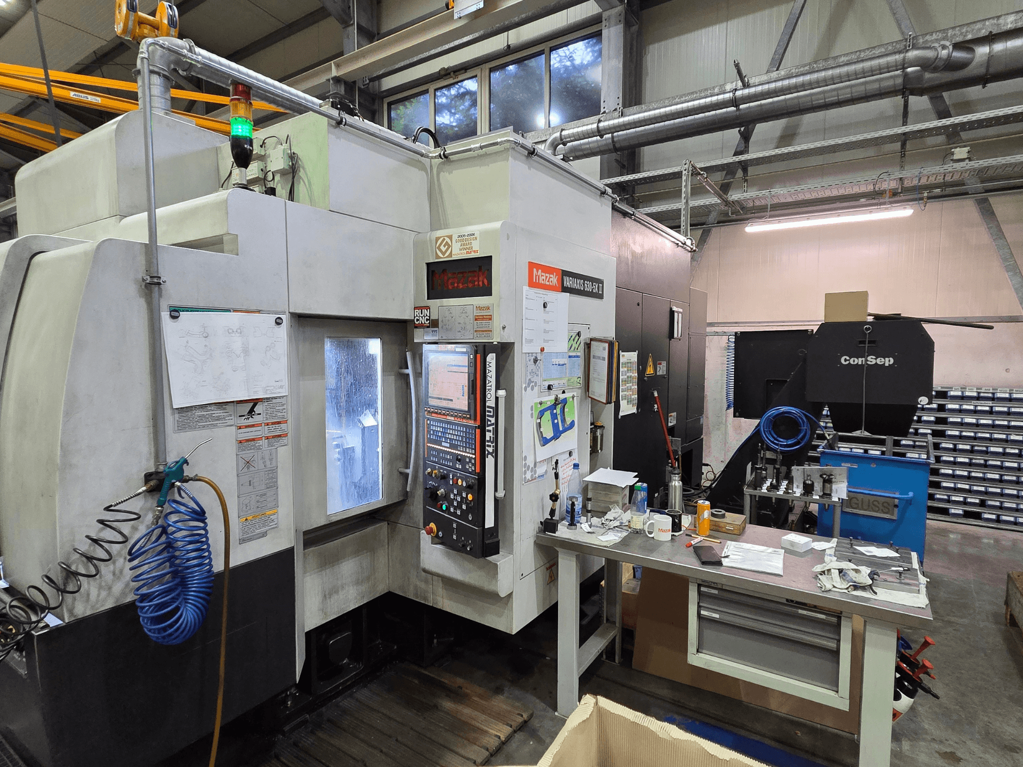 Máquina CNC Mazak Variaxis 630-5X numa oficina, vista frontal com painel de controlo, mangueira e acessórios na mesa lateral.
