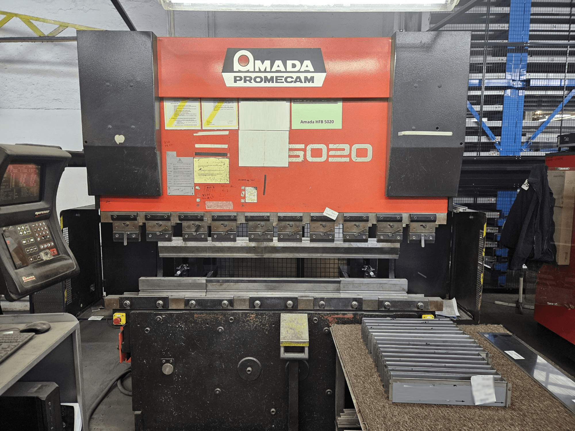 Prensa dobradeira Amada HFBO 5020 em vista frontal, com painel de controlo e peças metálicas empilhadas na superfície de trabalho.