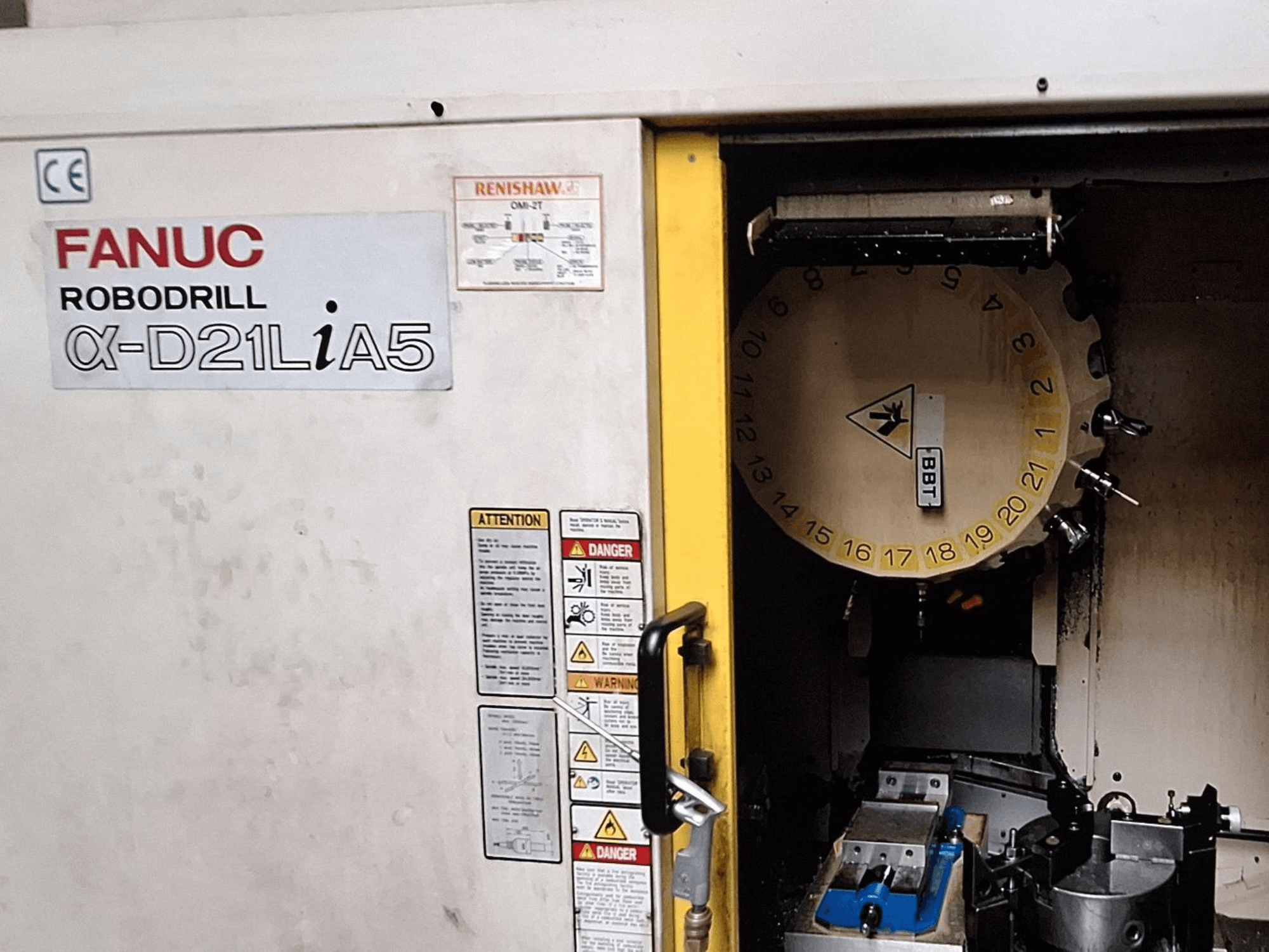 FANUC Robodrill α-D21LiA5, vista frontal. Mostra o carrossel de ferramentas e a área de maquinação com pinças e avisos na porta.