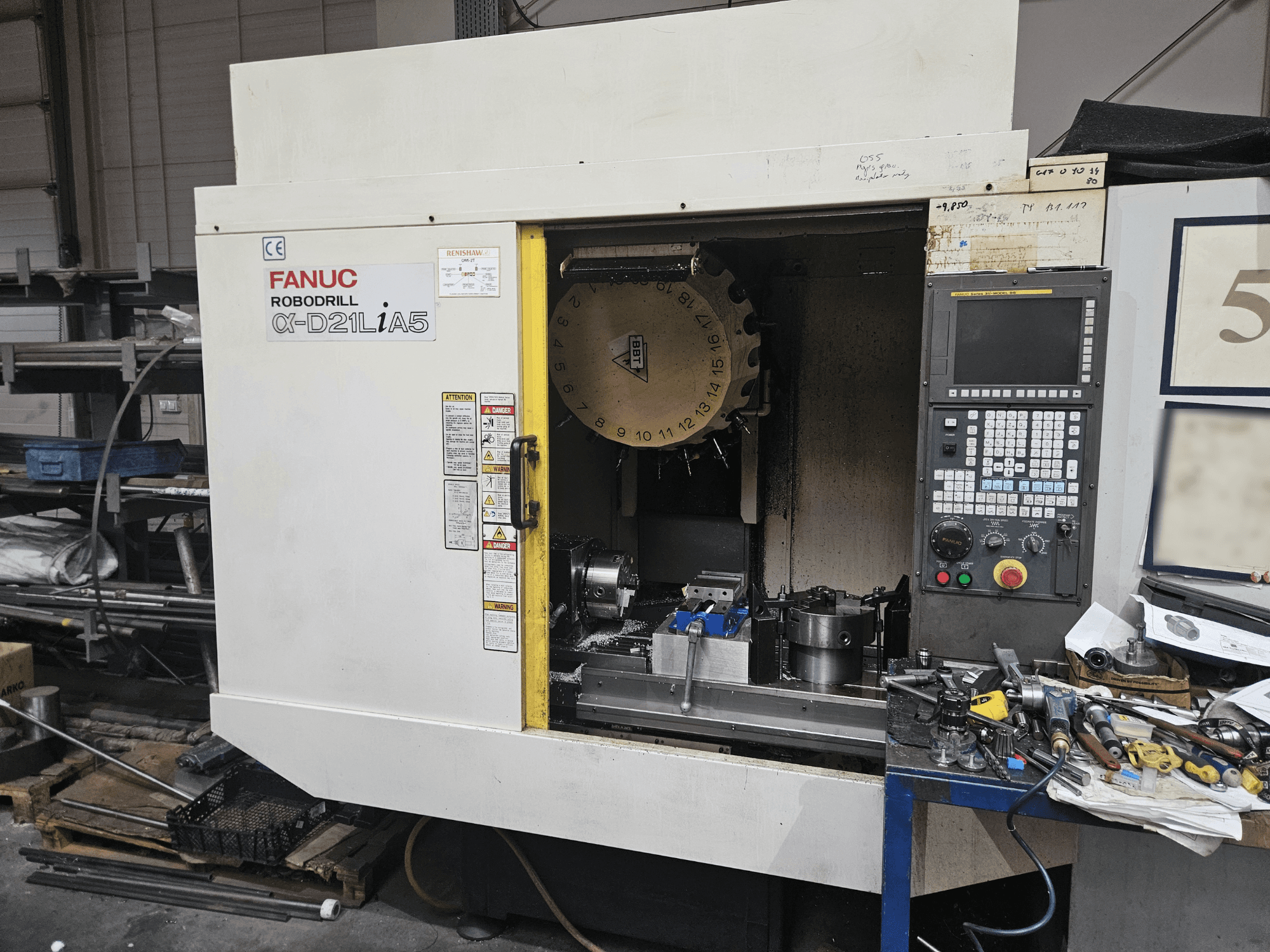 FANUC Robodrill α-D21LiA5, vista frontal mostrando o suporte de ferramentas e o painel de controlo, rodeado de ferramentas e equipamento numa oficina.