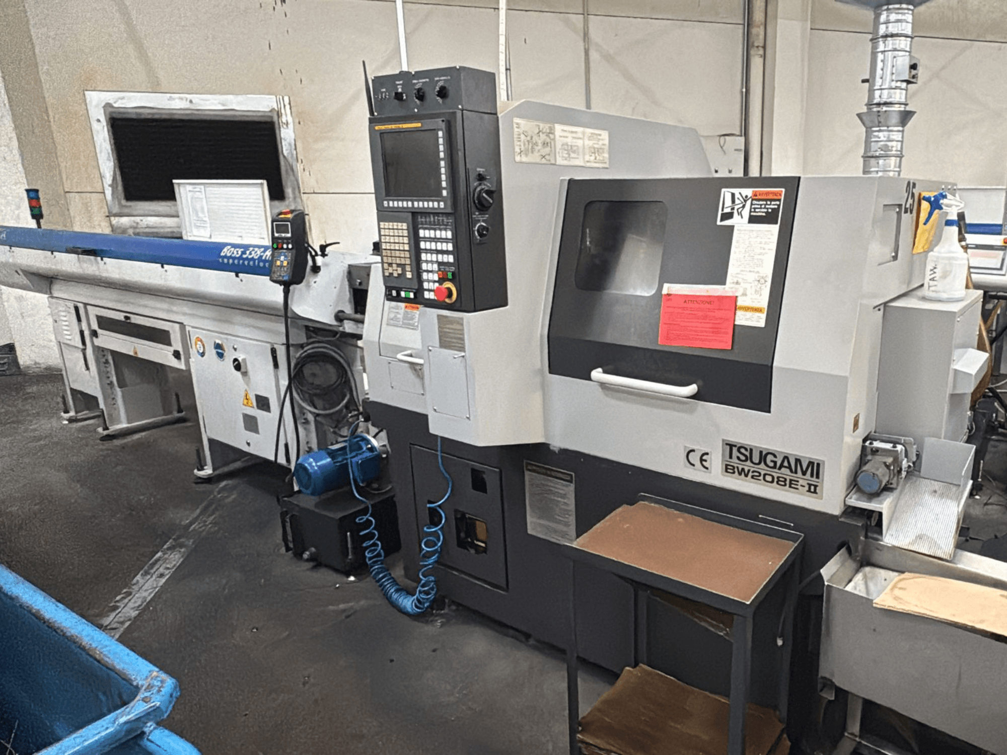 Máquina CNC Tsugami BW208E-II numa oficina, mostrada de um ângulo frontal, com o painel de controlo e as etiquetas de segurança visíveis.