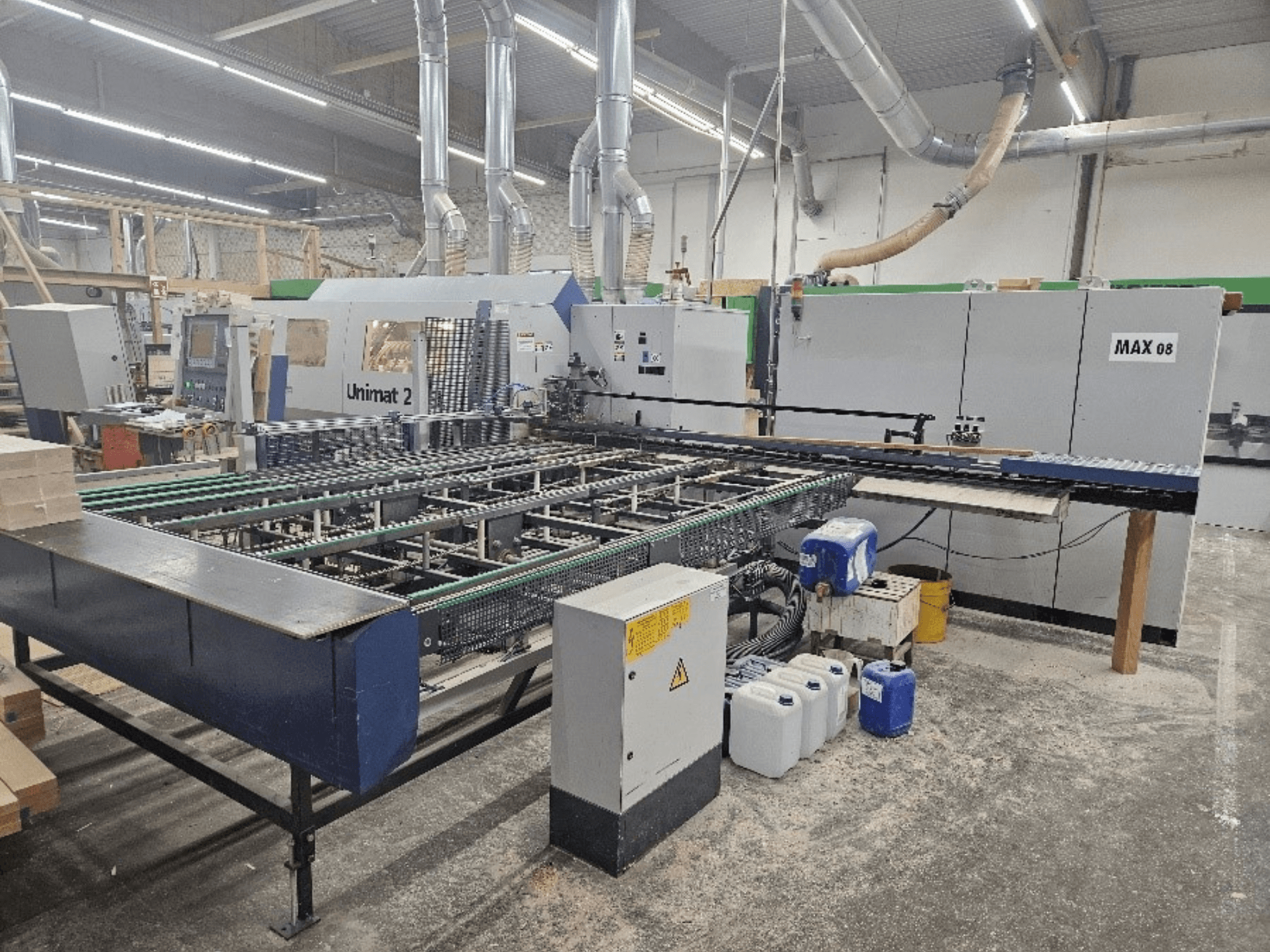 Vista lateral de uma máquina CNC Unimat 2 numa oficina, com correias transportadoras e componentes técnicos com a unidade Max 08 visível.