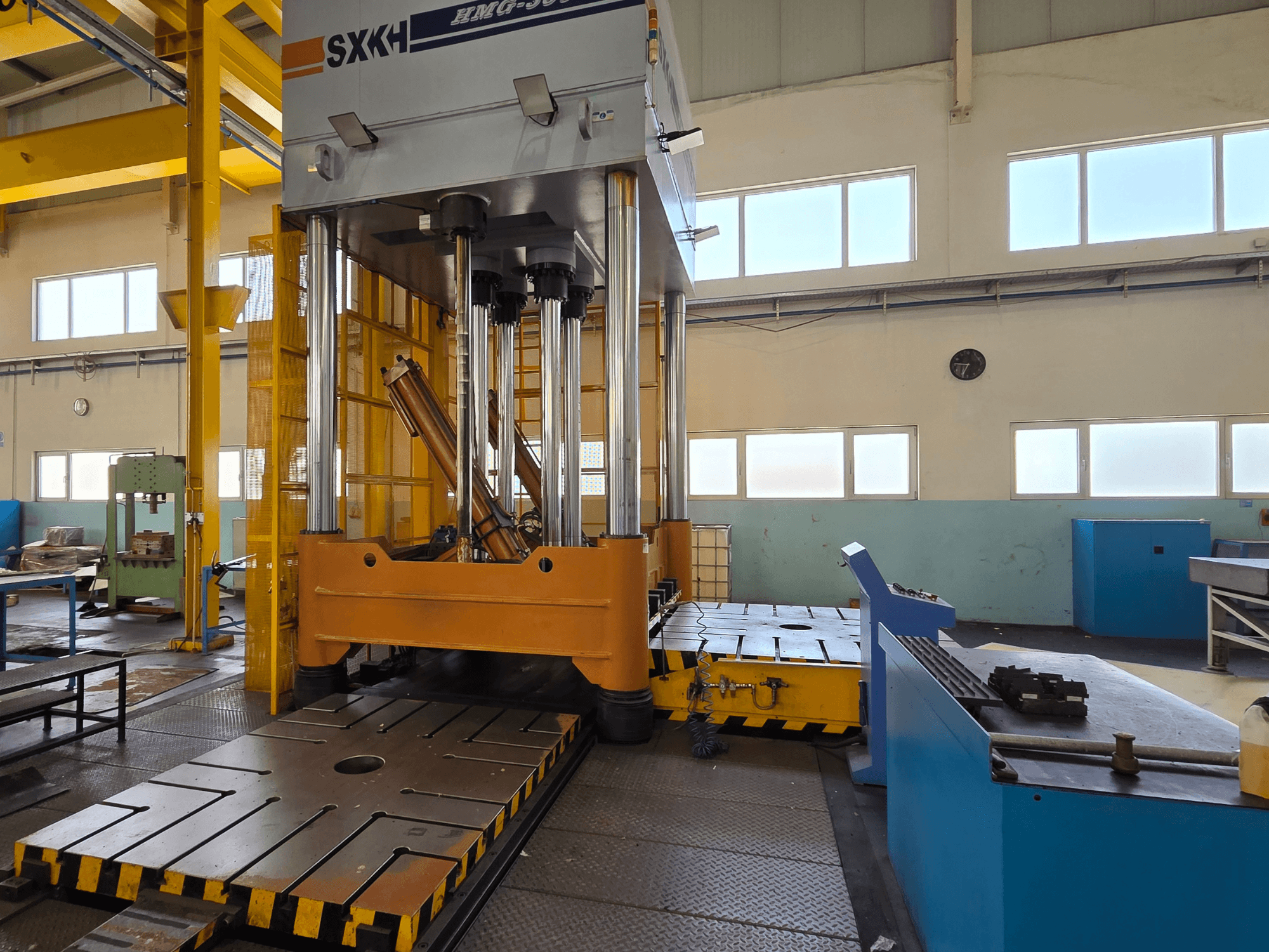 Vista frontal da prensa hidráulica SXKH HMG-300JM, com grandes cilindros de prensagem e uma base robusta numa fábrica.