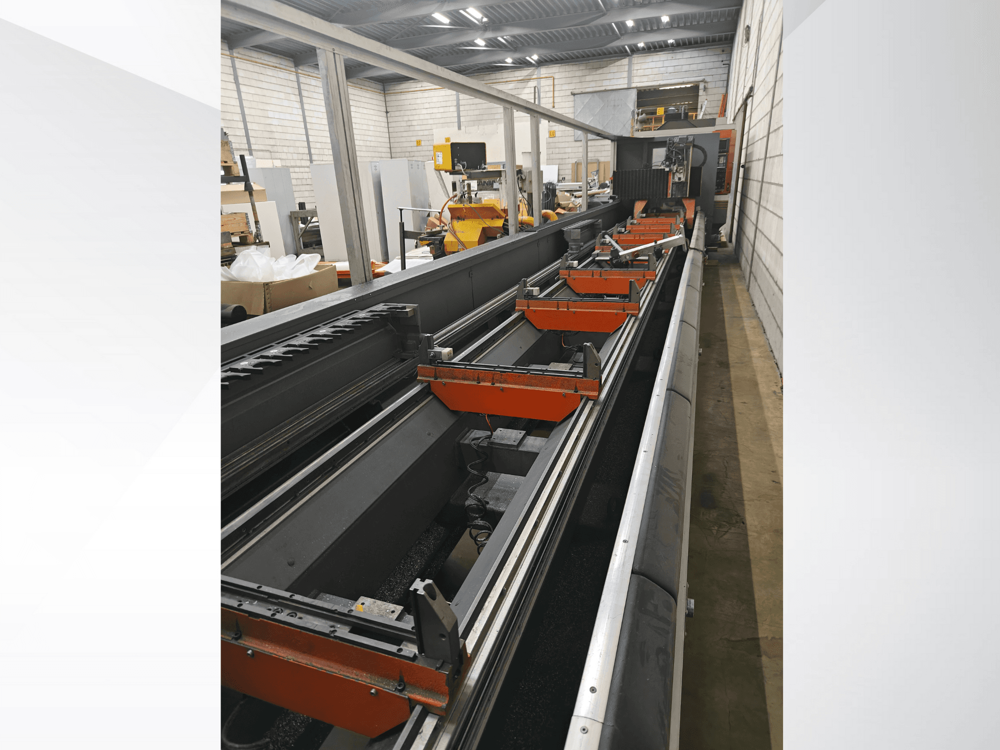 Vista lateral de uma máquina CNC industrial, com calhas lineares, pinças laranja e componentes de precisão num armazém.