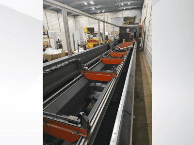 Vista lateral de uma máquina CNC industrial, com calhas lineares, pinças laranja e componentes de precisão num armazém.
