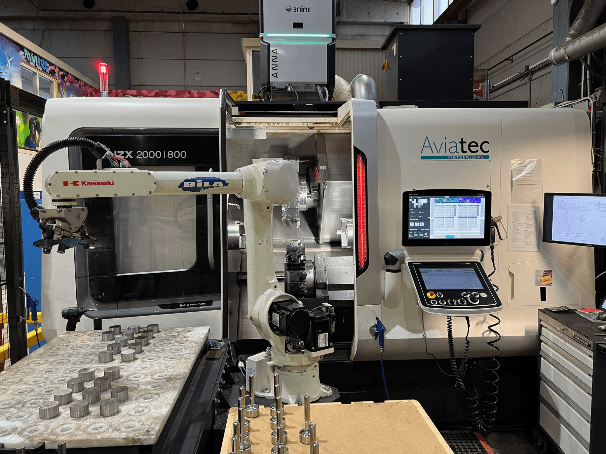 Torno horizontal DMG MORI NZX 2000/800SY com braço robótico Kawasaki, painel de controlo e componentes metálicos na mesa de trabalho.