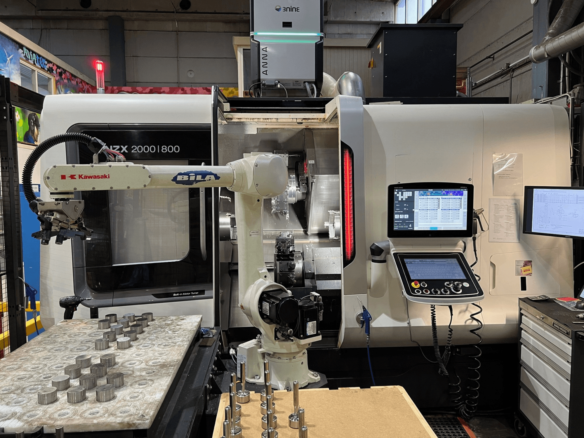 Torno horizontal DMG MORI NZX 2000/800SY numa oficina, com o braço robótico da Kawasaki e os comandos visíveis.