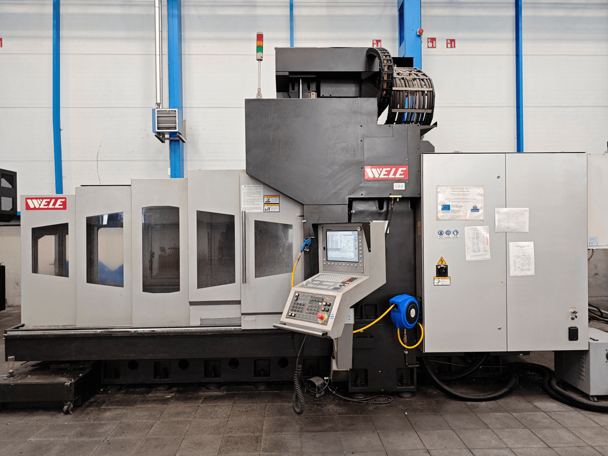 Centro de maquinação WELE em vista frontal, com painel de controlo, avisos de segurança e múltiplas janelas de acesso.