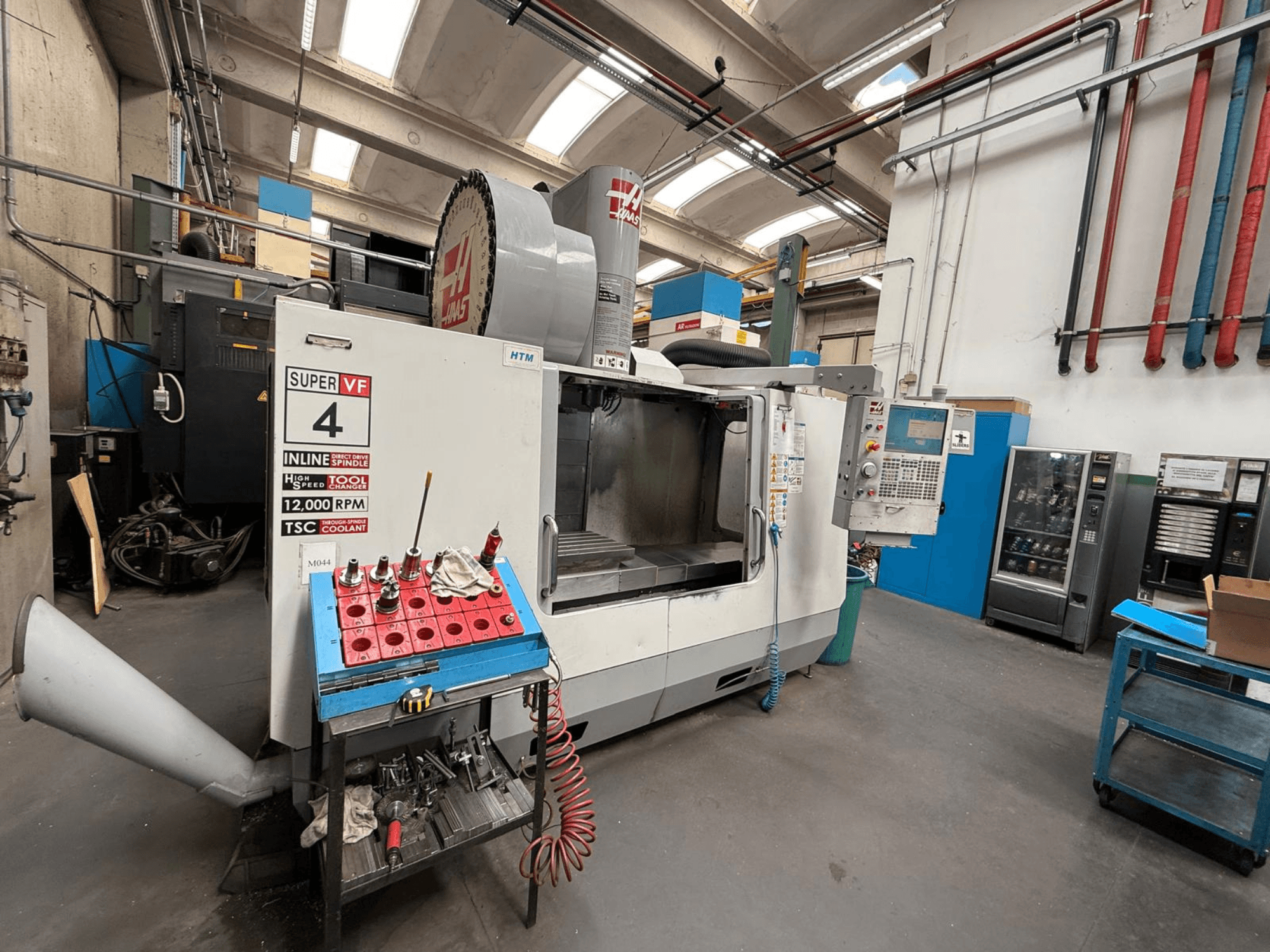 Fresadora CNC HAAS Super VF 4 com painel de controlo, fuso de 12.000 RPM e ferramentas numa prateleira de armazenamento azul, vista lateral.