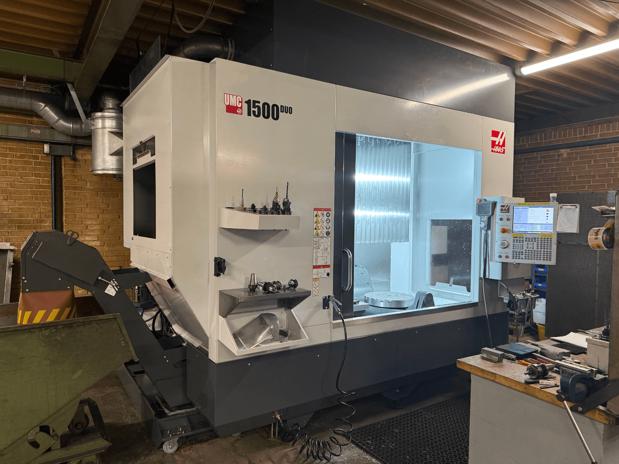 Máquina CNC UMC 1500DUO da Haas, vista frontal, apresenta uma grande janela, painel de controlo e suportes de ferramentas numa prateleira.