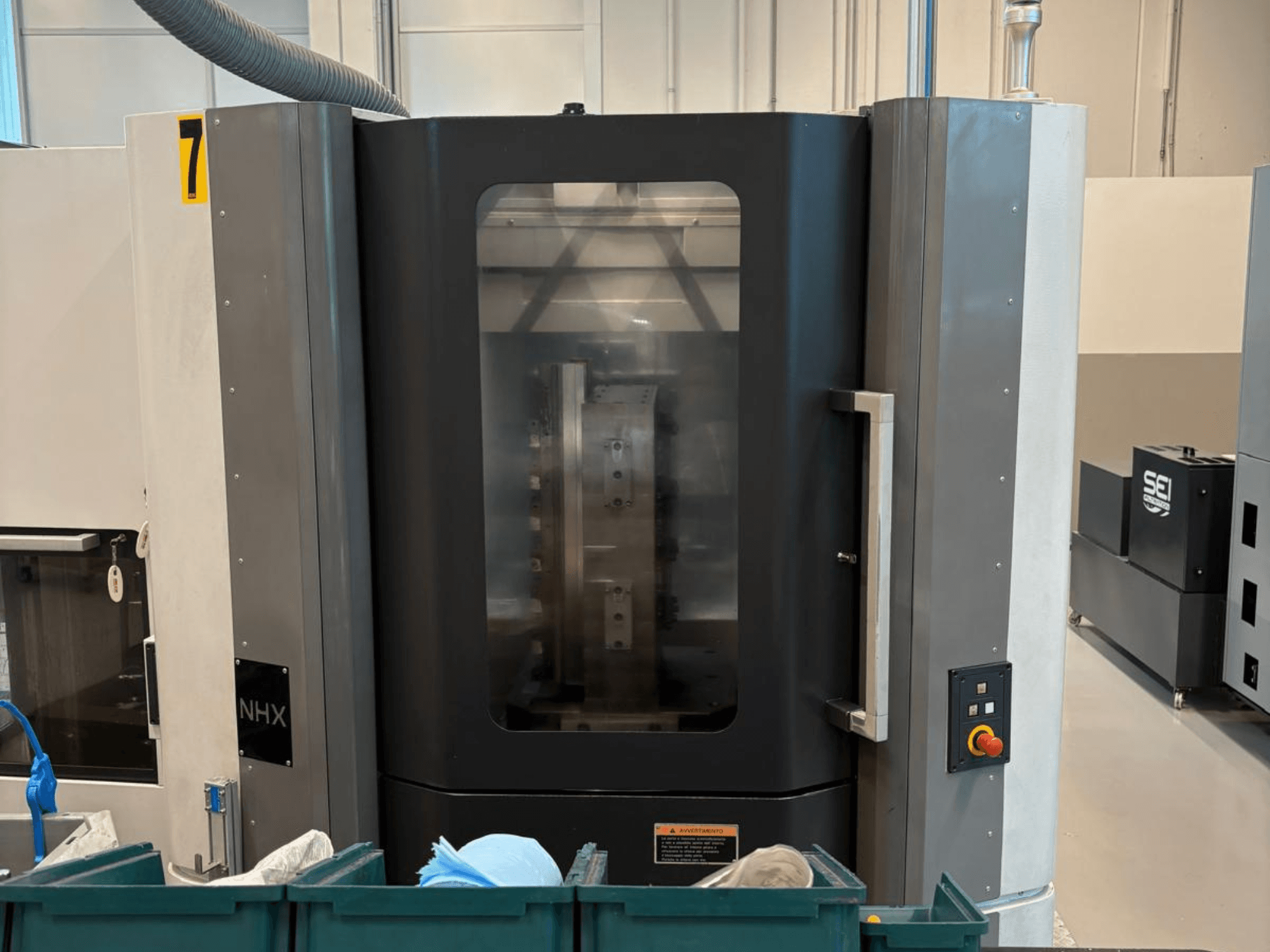 Máquina CNC NHX em vista frontal, mostrando o painel de controlo e o visor transparente; localizada numa oficina com caixotes verdes nas proximidades.