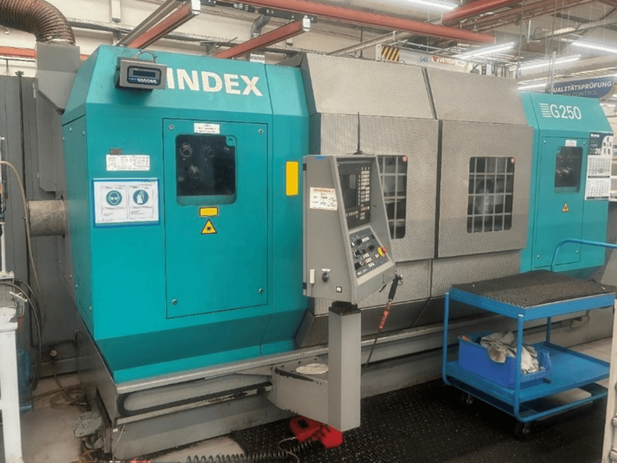 Torno horizontal INDEX G250, vista frontal que mostra o painel de controlo e a estrutura metálica, com etiquetas de segurança e um carrinho azul.