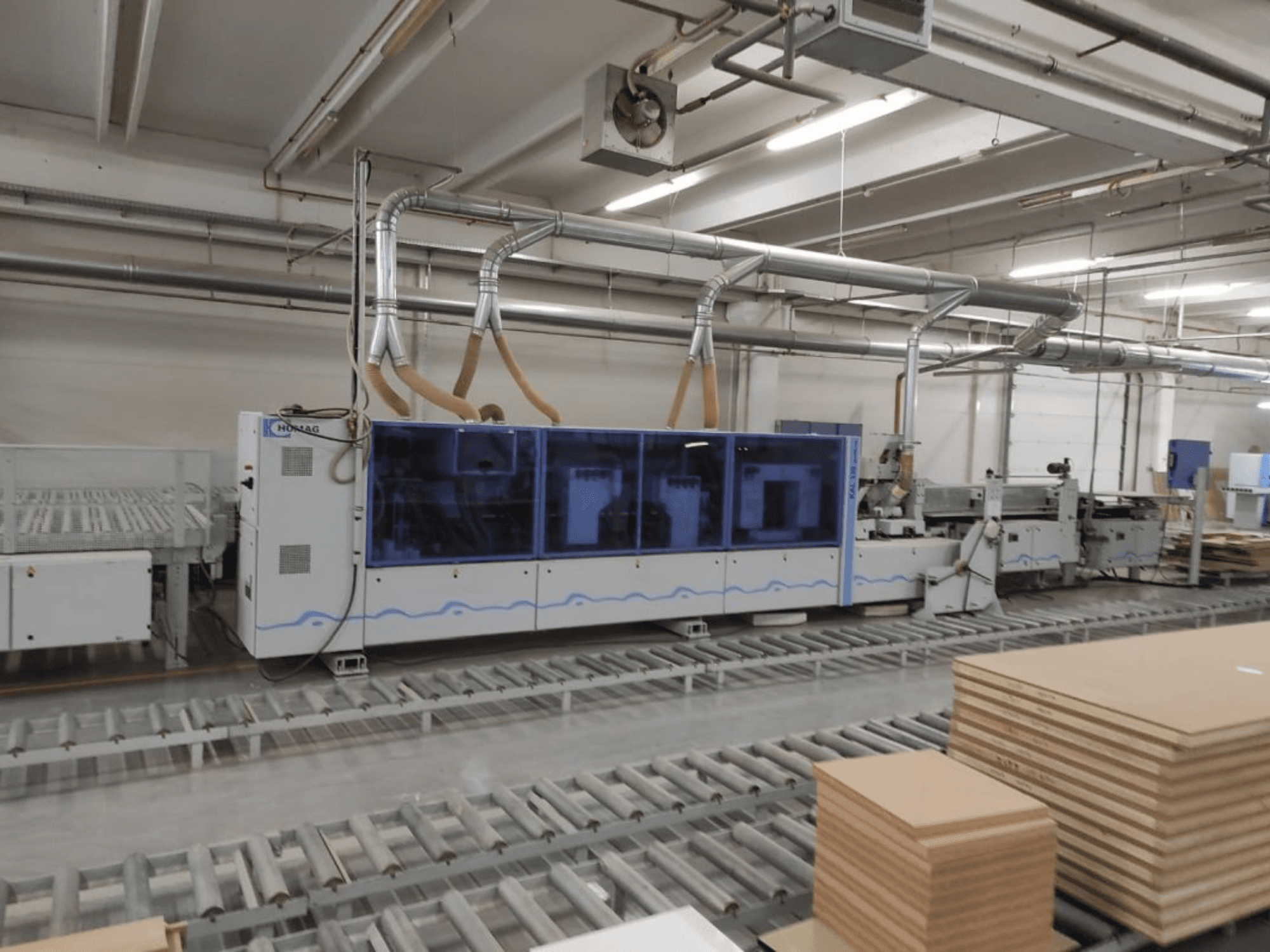 Máquina de produção HOMAG numa fábrica, vista frontal, com caixa azul e sistema de transporte com folhas de contraplacado empilhadas.
