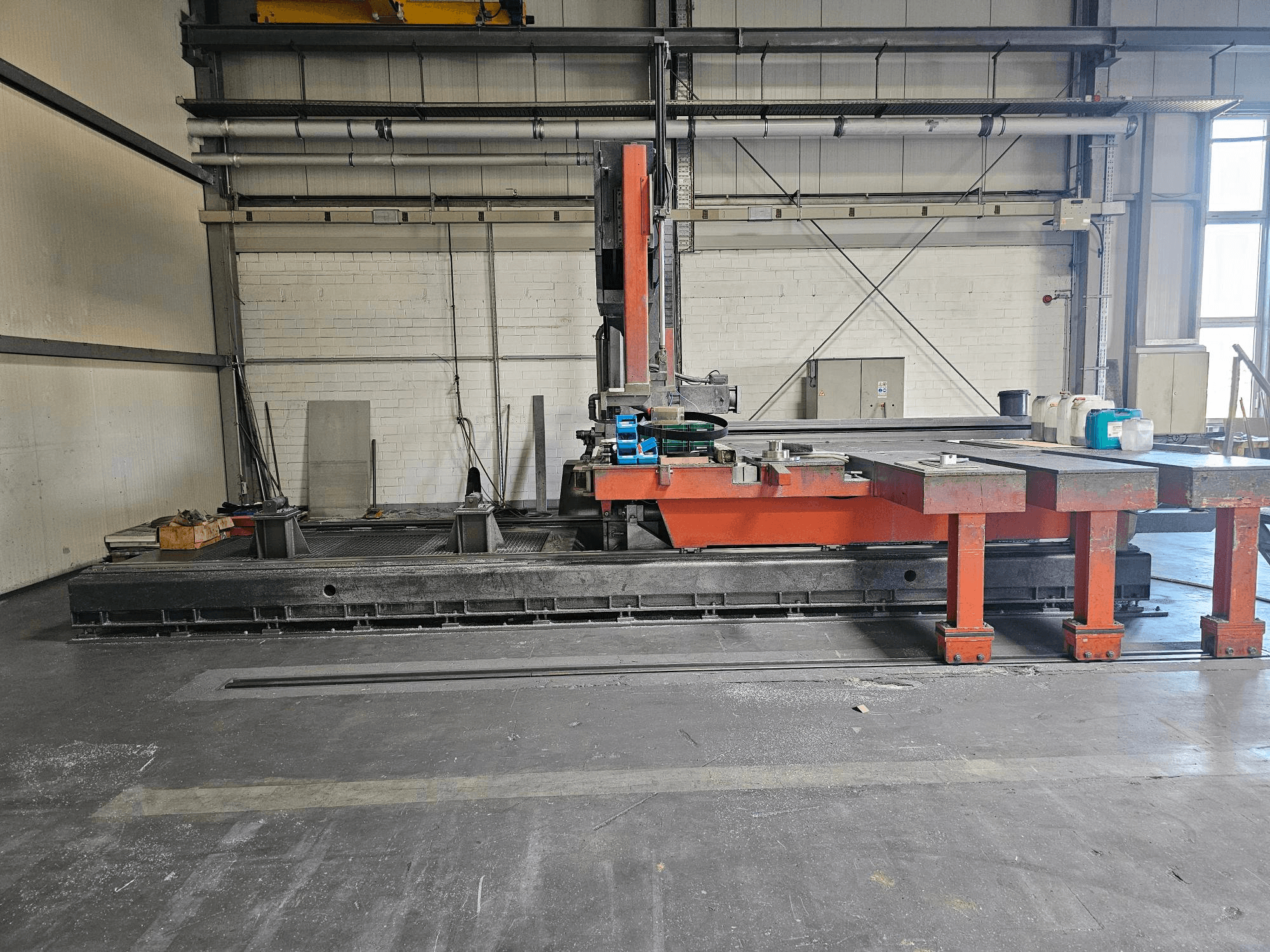 Máquina de corte industrial numa oficina aberta, cor de laranja e preta, com uma mesa grande; vista lateral mostrando os acessórios e o espaço de trabalho.