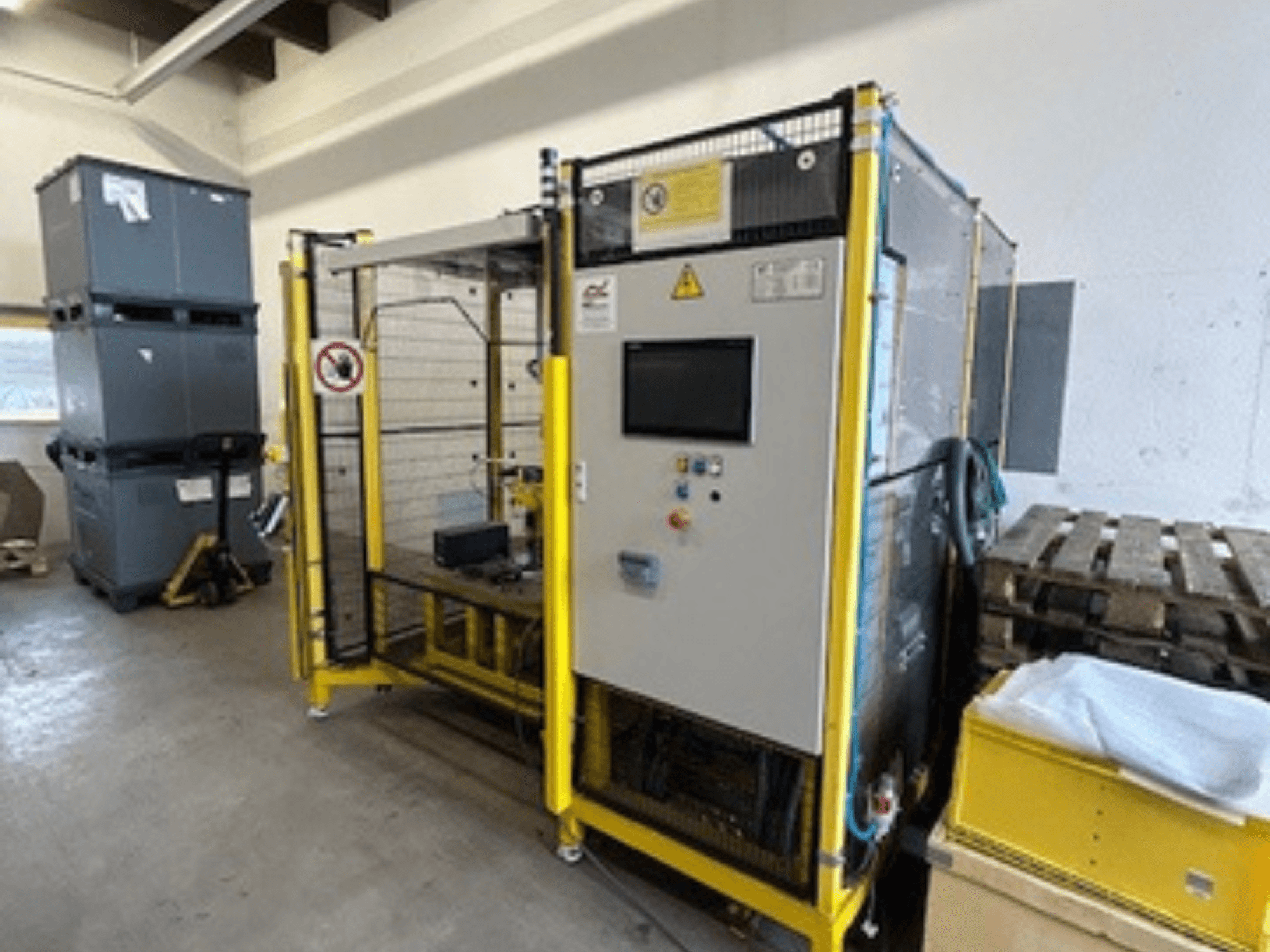 Máquina industrial com painel de controlo e dispositivos de segurança, vista frontal; quadro amarelo, caixas cinzentas e paletes em segundo plano.