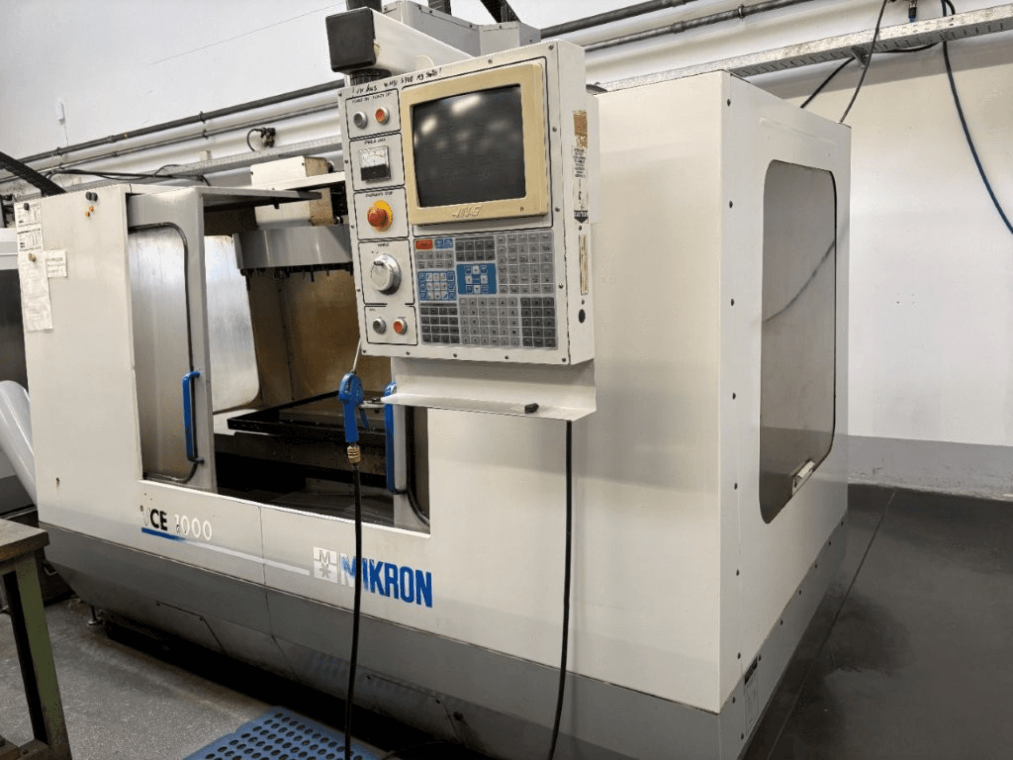 Máquina CNC Mikron CE 2000, vista frontal; apresenta um painel de controlo com botões e ecrã, exterior prateado e área de trabalho.