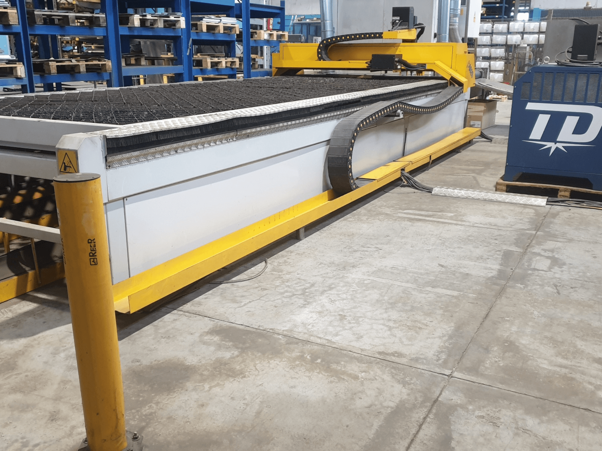 Máquina de corte industrial com acabamento amarelo, vista frontal, com uma base texturada e um painel de controlo, colocada no chão de uma fábrica.