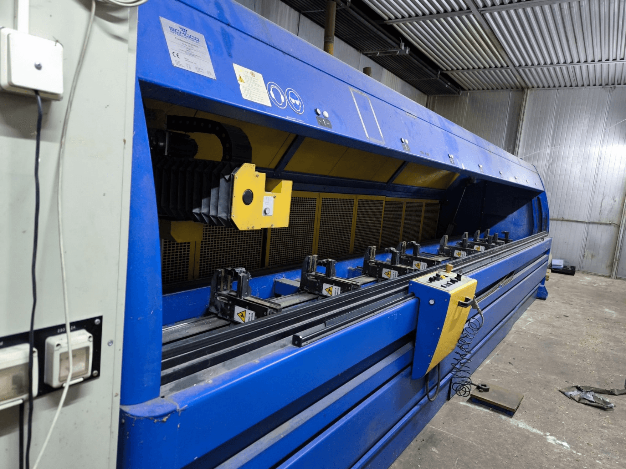 Máquina de corte industrial Schneider em azul, vista lateral, com painel de controlo, correias e várias estações de fixação visíveis.