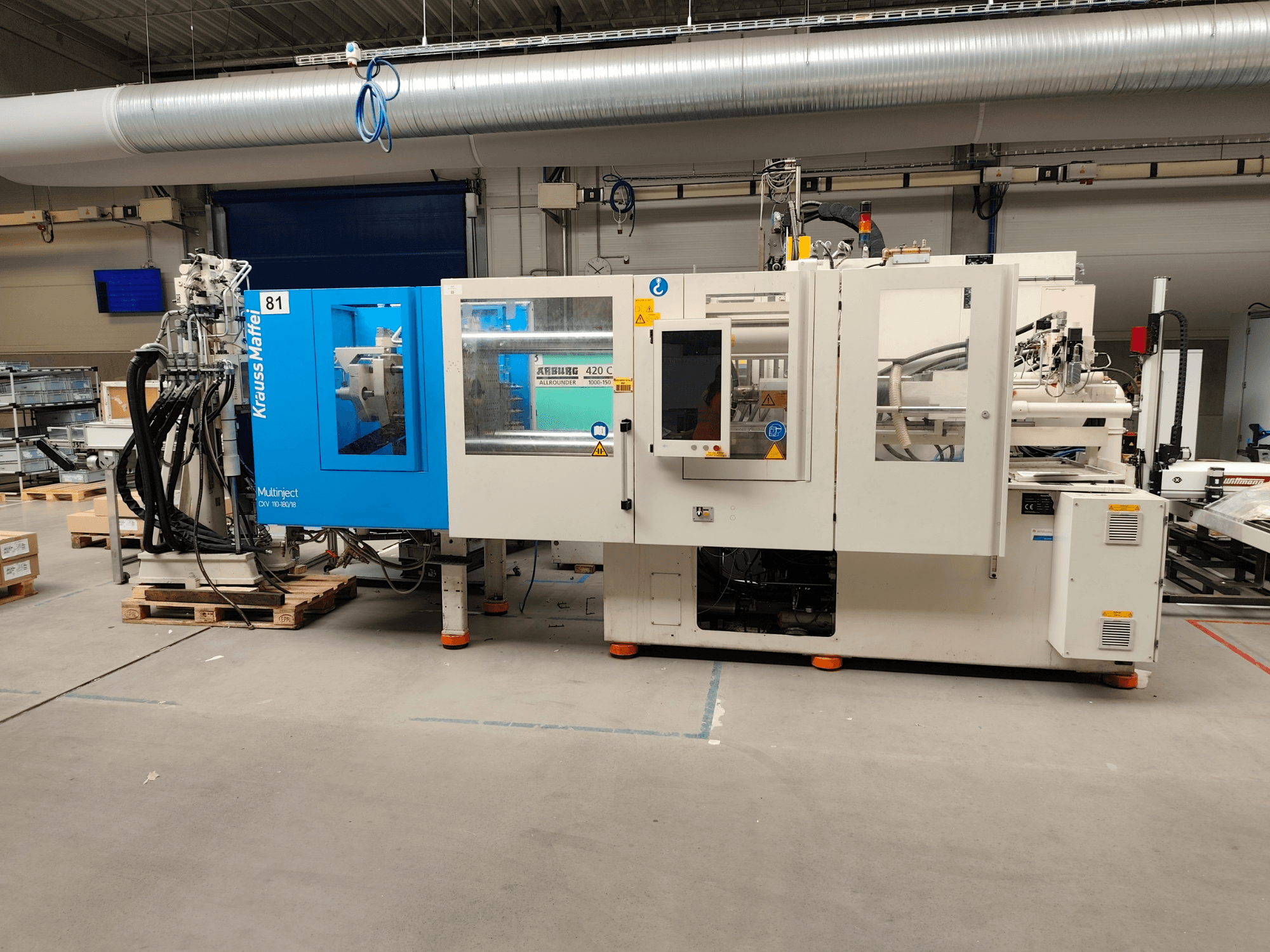 Máquina de moldagem por injeção hidráulica Krauss Maffei 110-180-180 CX V, vista lateral, mostrando a estrutura azul e branca, o painel de controlo e os conectores.