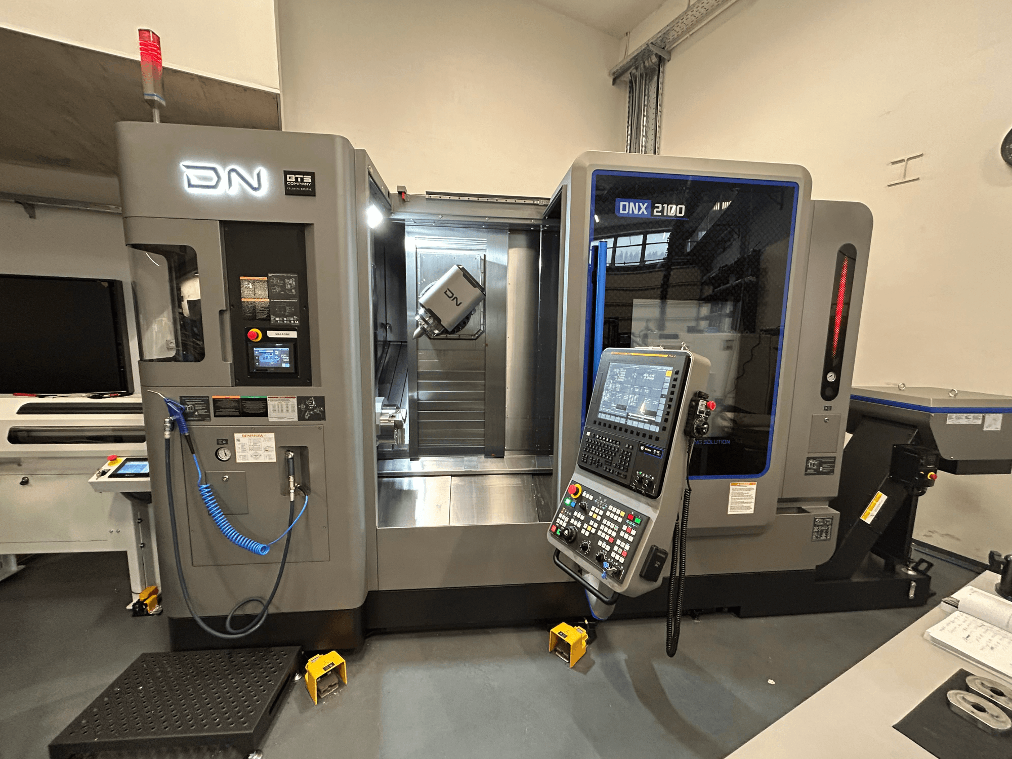 Máquina CNC DN ONX 2100, vista frontal, com painel de controlo, área de trabalho e ferramenta. Equipada com luz de segurança e indicadores de funcionamento.
