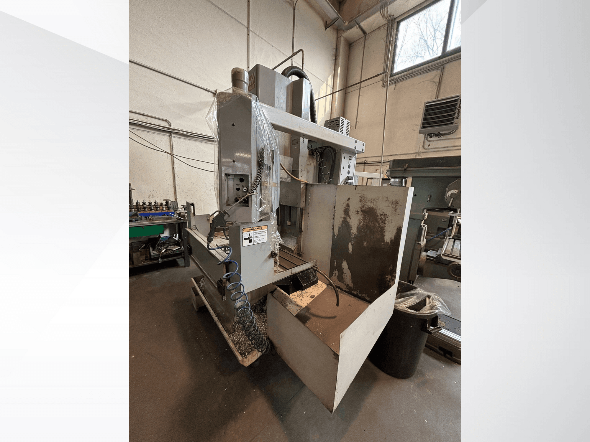 Fresadora vertical com cobertura de proteção, vista lateral; inclui mangueira de refrigeração e painel de controlo visível. Ideal para maquinagem.