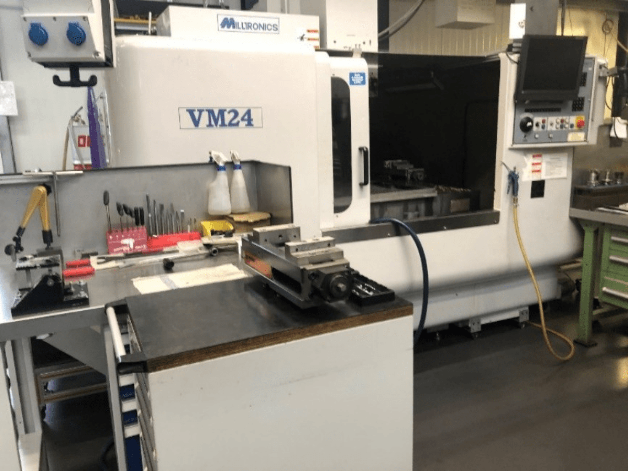 Espaço de trabalho de maquinaria com a máquina CNC Milltronics VM24. Vista frontal com ferramentas e equipamento numa bancada de trabalho.