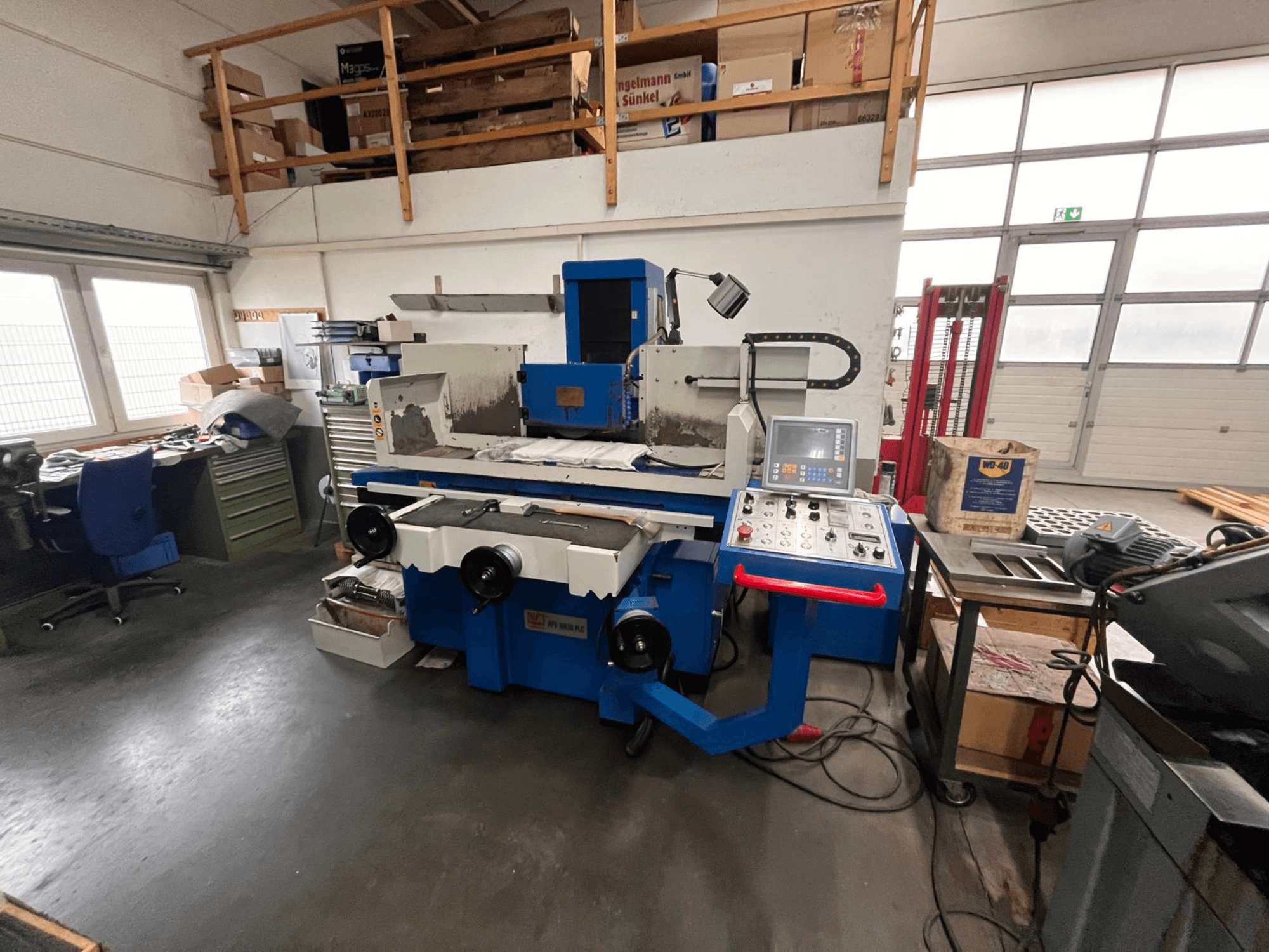 Esmeriladora de superfície azul com painel de controlo digital, vista frontal, mostrando a desarrumação do espaço de trabalho e um armário de ferramentas verde.