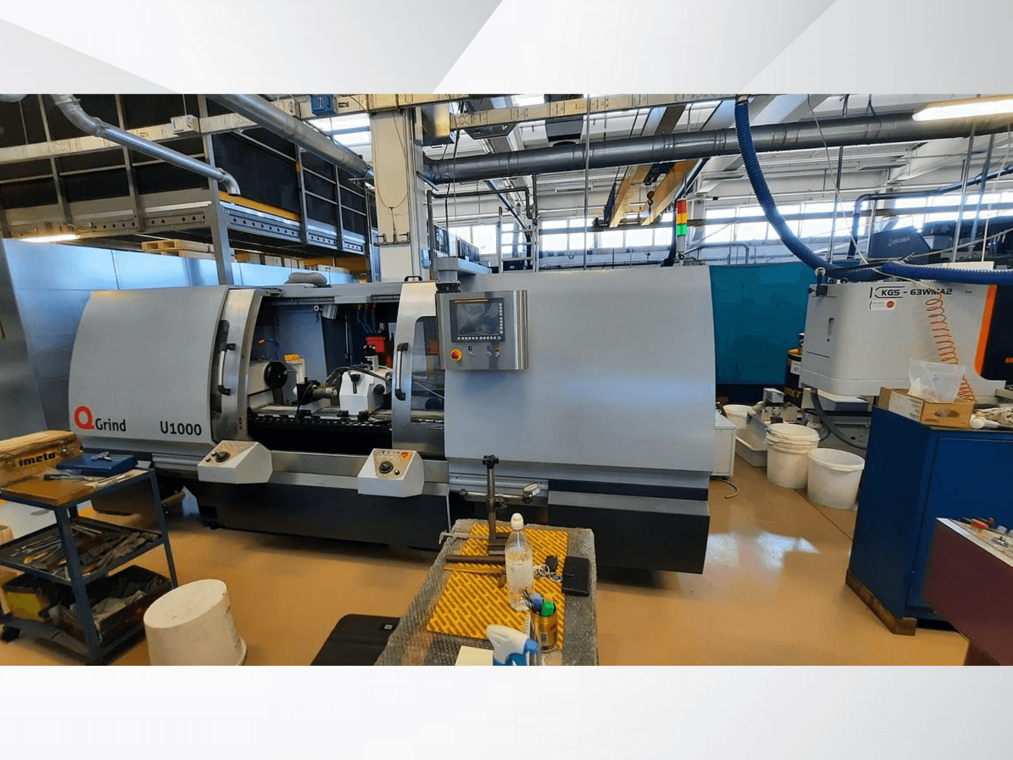Máquina Q-Grind U1000 numa oficina, mostrando uma vista frontal com painel de controlo e área de corte aberta, rodeada de equipamento.