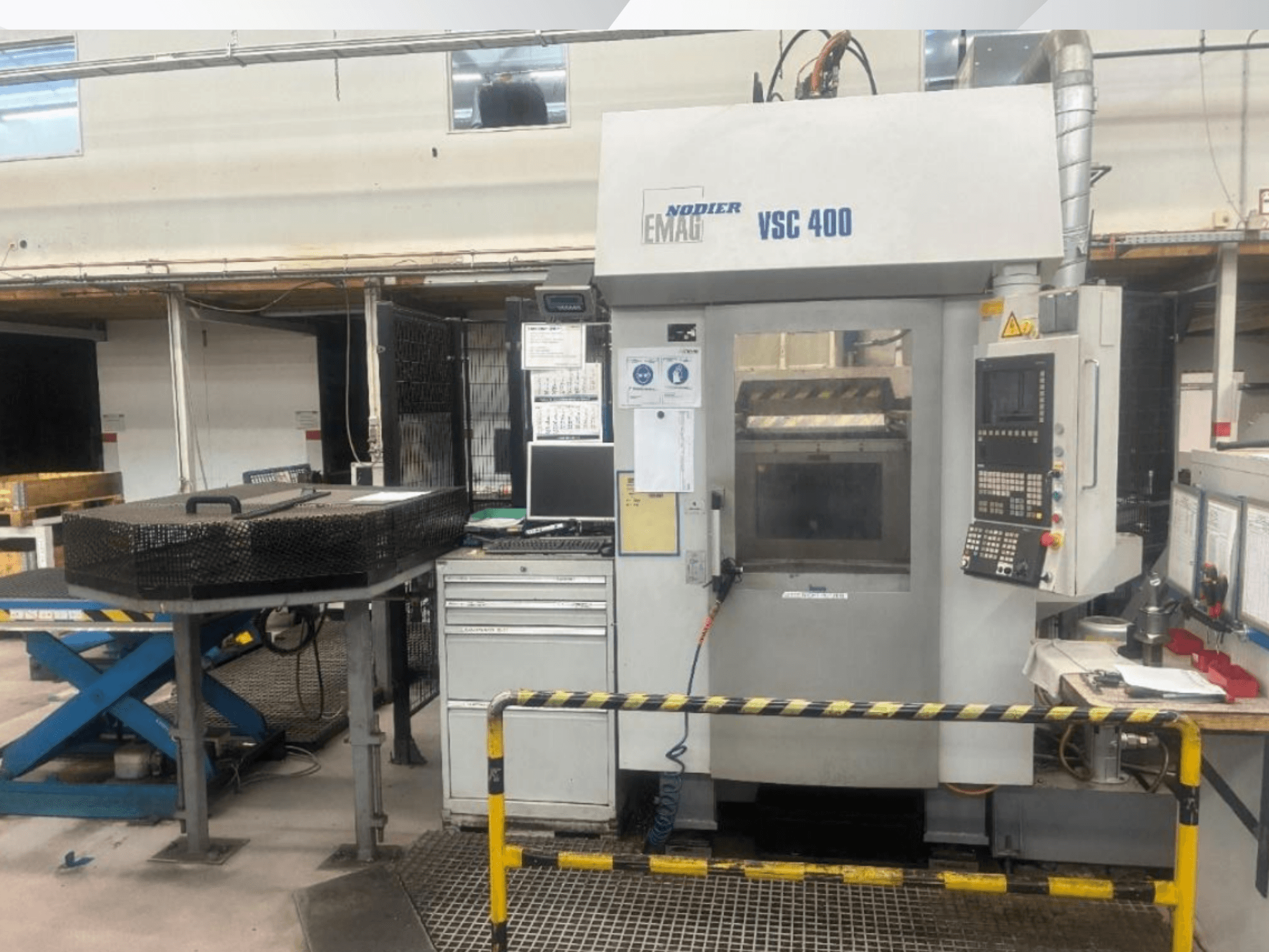 Vista frontal da máquina de torneamento vertical EMAG VSC 400, com destaque para o painel de controlo, as barreiras de segurança e o equipamento circundante, numa oficina bem iluminada.