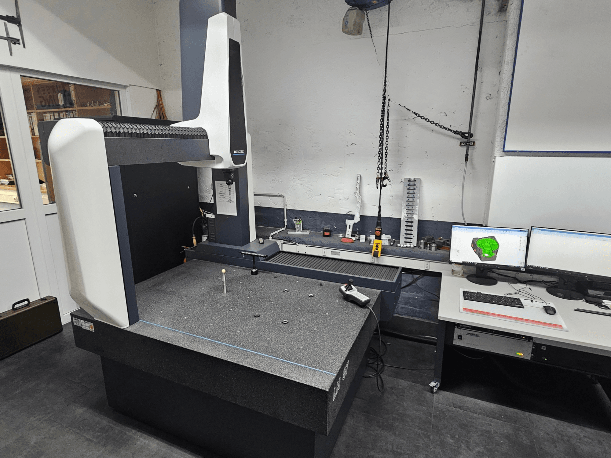 Máquina de medição por coordenadas Wenzel LH 87 numa oficina, vista frontal, com um apalpador na mesa de granito e dois monitores a mostrar o desenho CAD.
