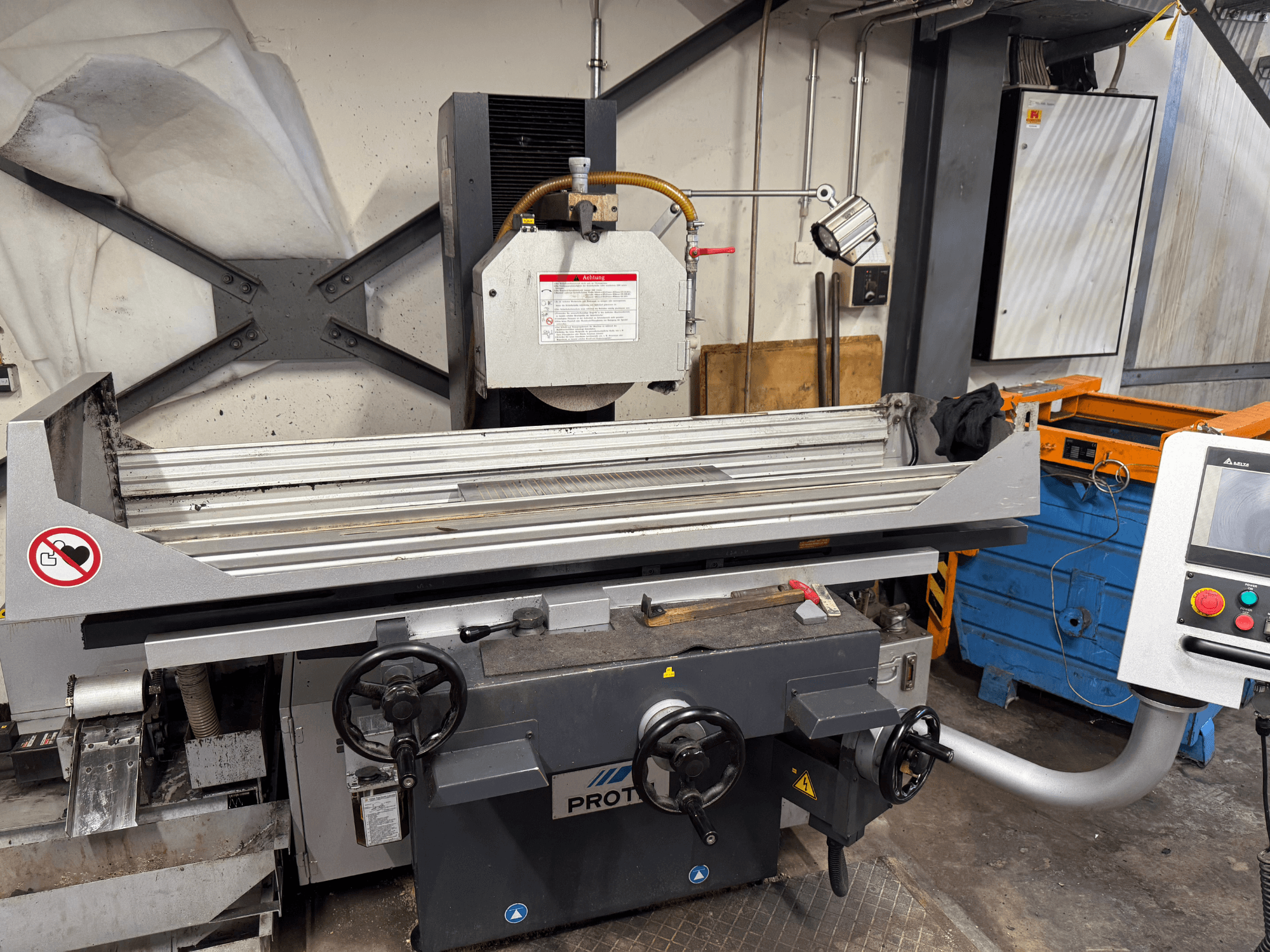 Máquina PROTEC, vista frontal, mostrando uma rebarbadora de superfície com comandos manuais, avisos de segurança e um sistema de arrefecimento.