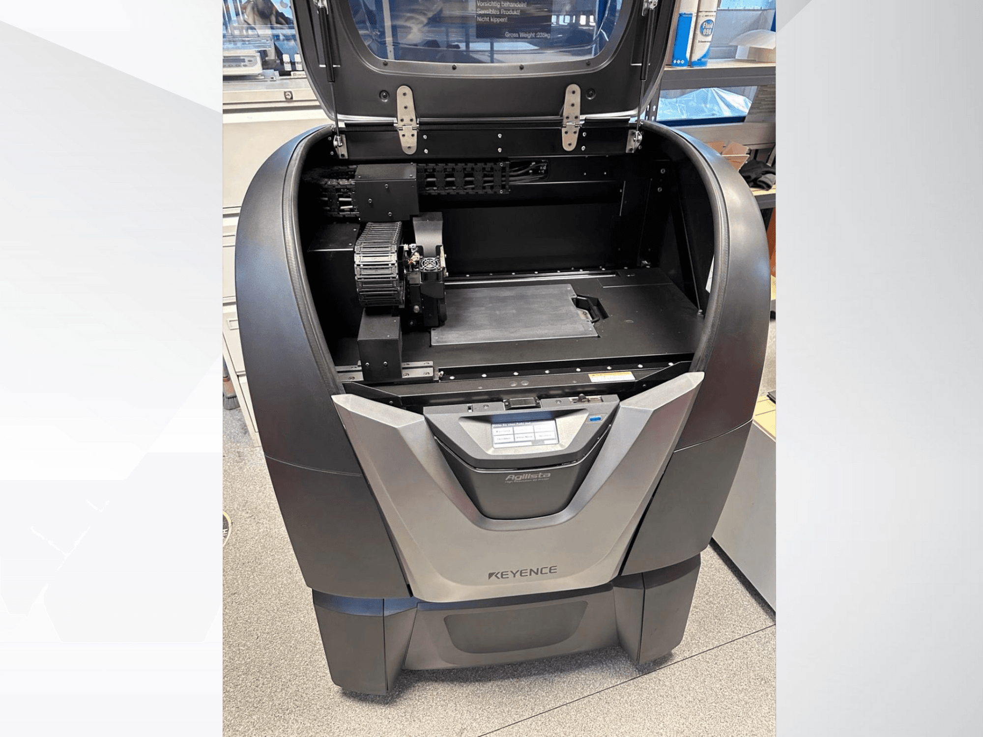 Impressora 3D KEYENCE Agilista, vista frontal, revela a parte superior aberta, a grande plataforma de construção e os componentes mecânicos internos.