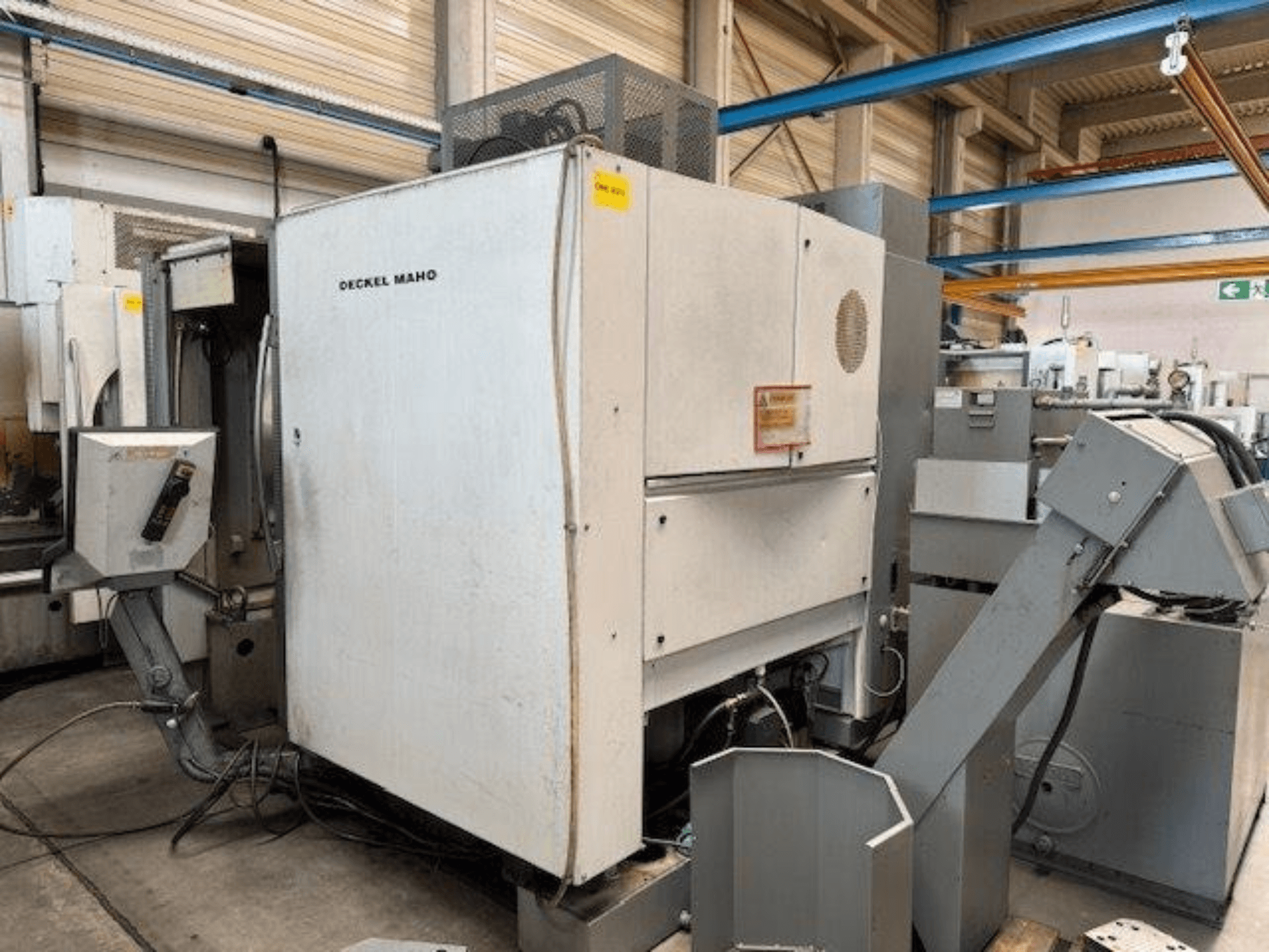 Centro de maquinação vertical DECKEL MAHO DMC 100 V, vista lateral com painel de controlo e vários acessórios visíveis.