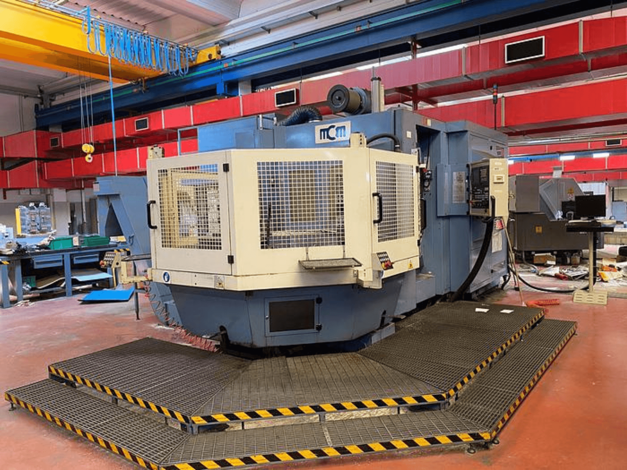 Máquina CNC MCM numa fábrica, vista frontal com gaiola de proteção, rebordos de segurança pretos e amarelos, rodeada por equipamento industrial.