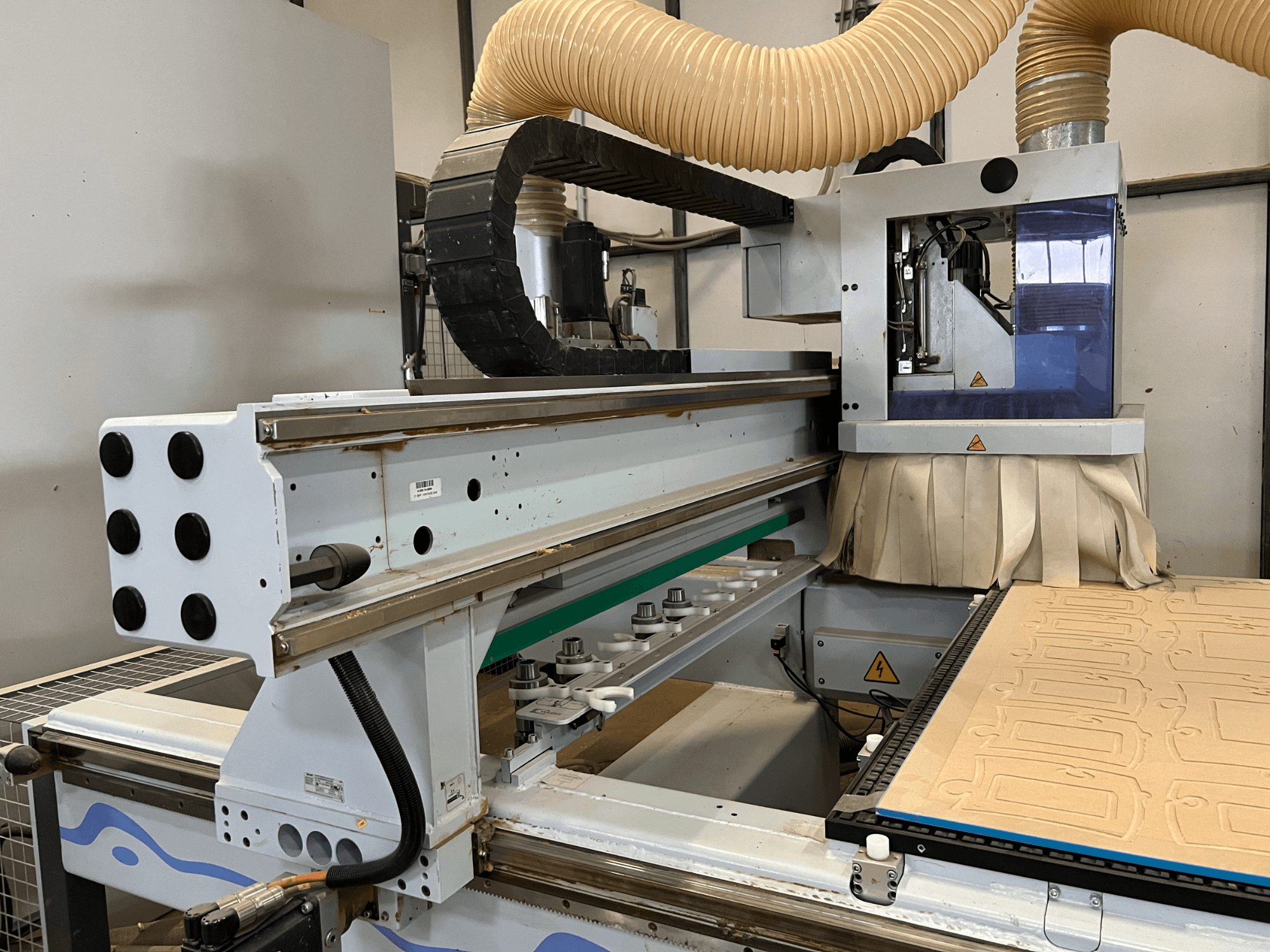 Vista lateral de uma fresadora CNC com um painel de controlo azul e prateado, equipada com extração de poeiras e uma mesa de corte de madeira.