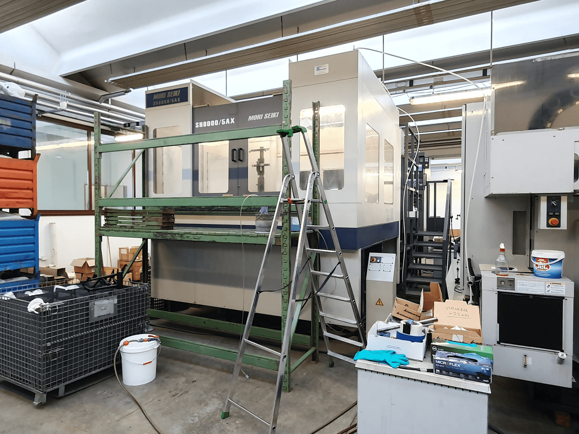 Máquina CNC Mori Seiki SH-8000/5AX numa oficina, vista frontal, com ferramentas de oficina e caixas de arrumação nas proximidades.