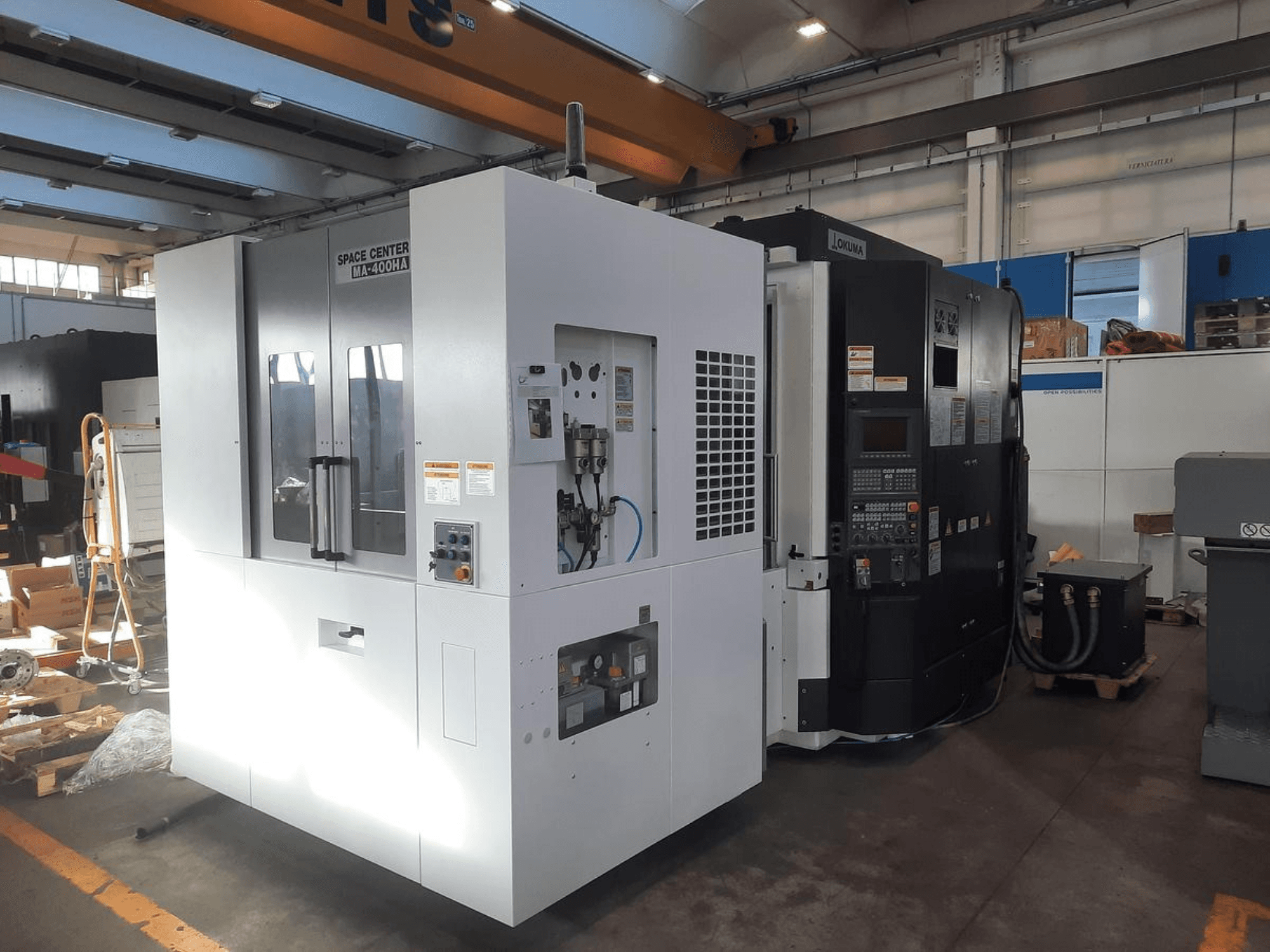 Vista frontal das máquinas CNC SPACE CENTER IM-400HAA e OKUMA, mostrando vários controlos e etiquetas do fabricante.