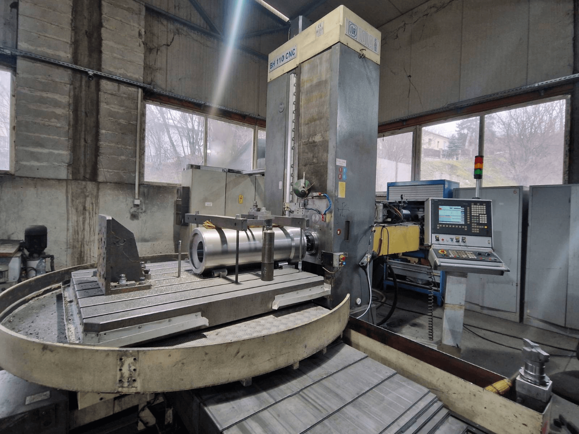 Máquina de furar CNC LOLA BH 110 numa oficina, vista frontal, com peça de trabalho em metal e painel de controlo.