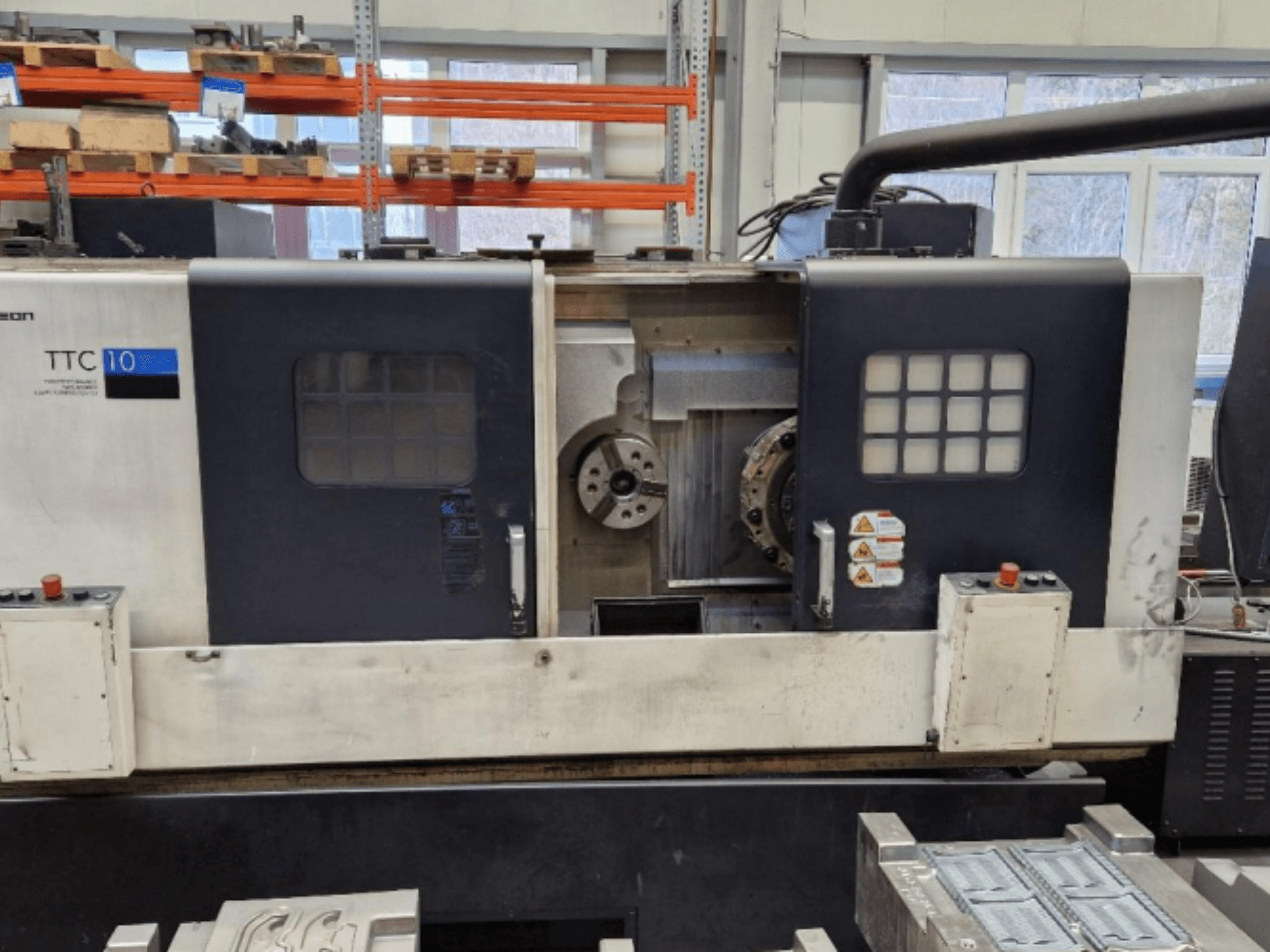 Vista frontal da máquina de torno TTC 10, mostrando o painel de controlo, mandris visíveis e espaço de trabalho com peças metálicas.