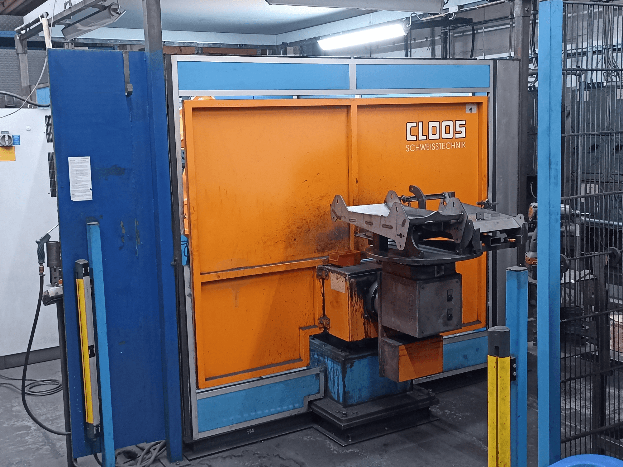 Robô de soldadura CLOOS Romat 320 numa oficina, vista frontal, com um painel de controlo laranja e uma peça metálica numa base rotativa.