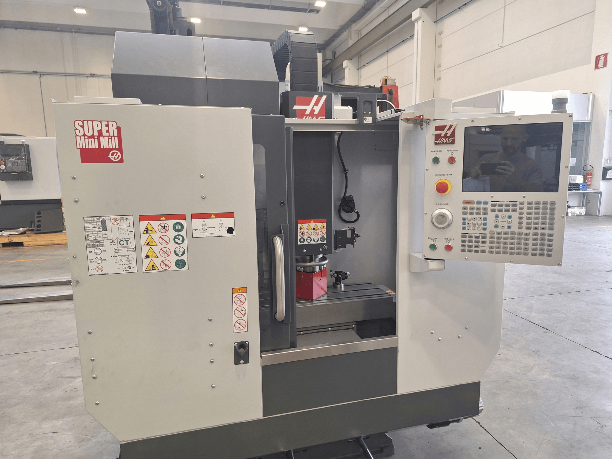 Vista frontal da Super Mini Mill da HAAS, mostrando o painel de controlo, as etiquetas de segurança e a área de maquinagem com uma peça montada.