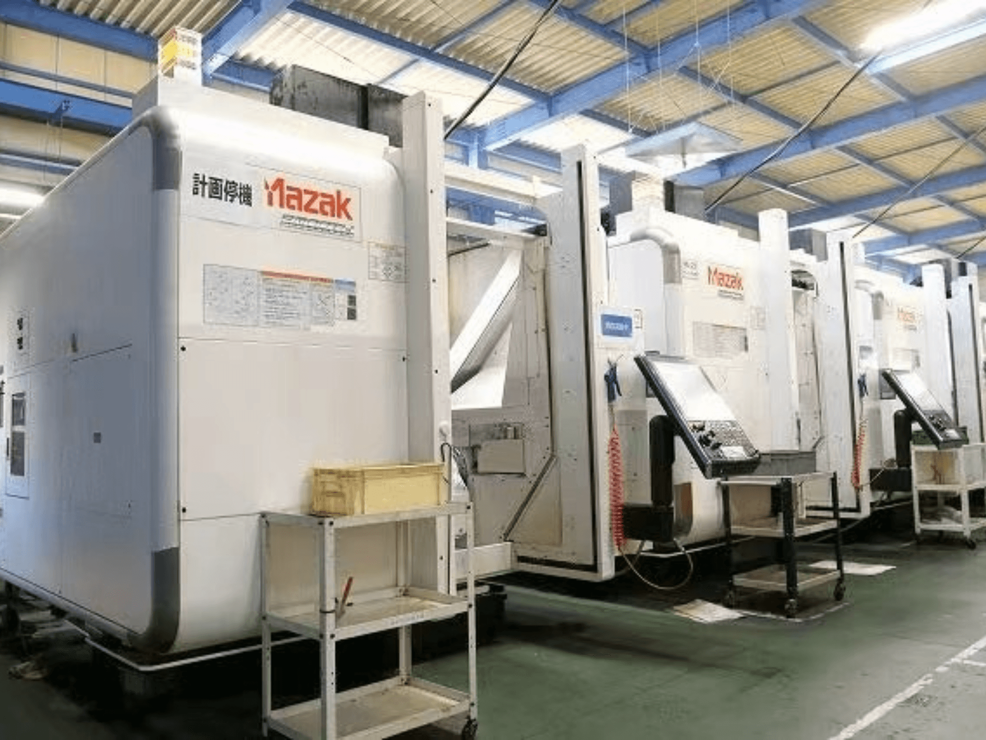 Máquinas CNC Mazak numa fábrica, vista lateral mostrando painéis de controlo e etiquetas em corpos brancos.