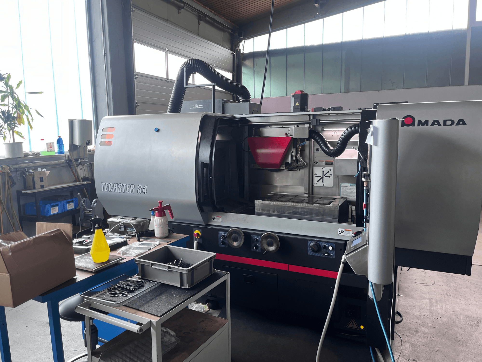 Máquina de retificação plana AMADA Techster 84 numa oficina, apresentando uma vista frontal com os comandos operacionais e o funil vermelho.