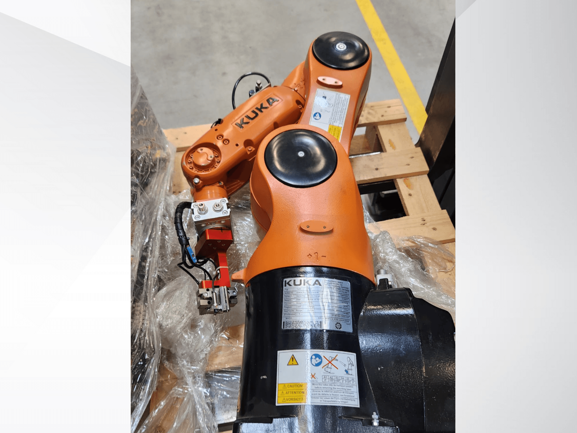 Braço robótico KUKA com acabamento laranja, vista lateral, com uma pinça e cablagem de controlo numa palete de madeira.